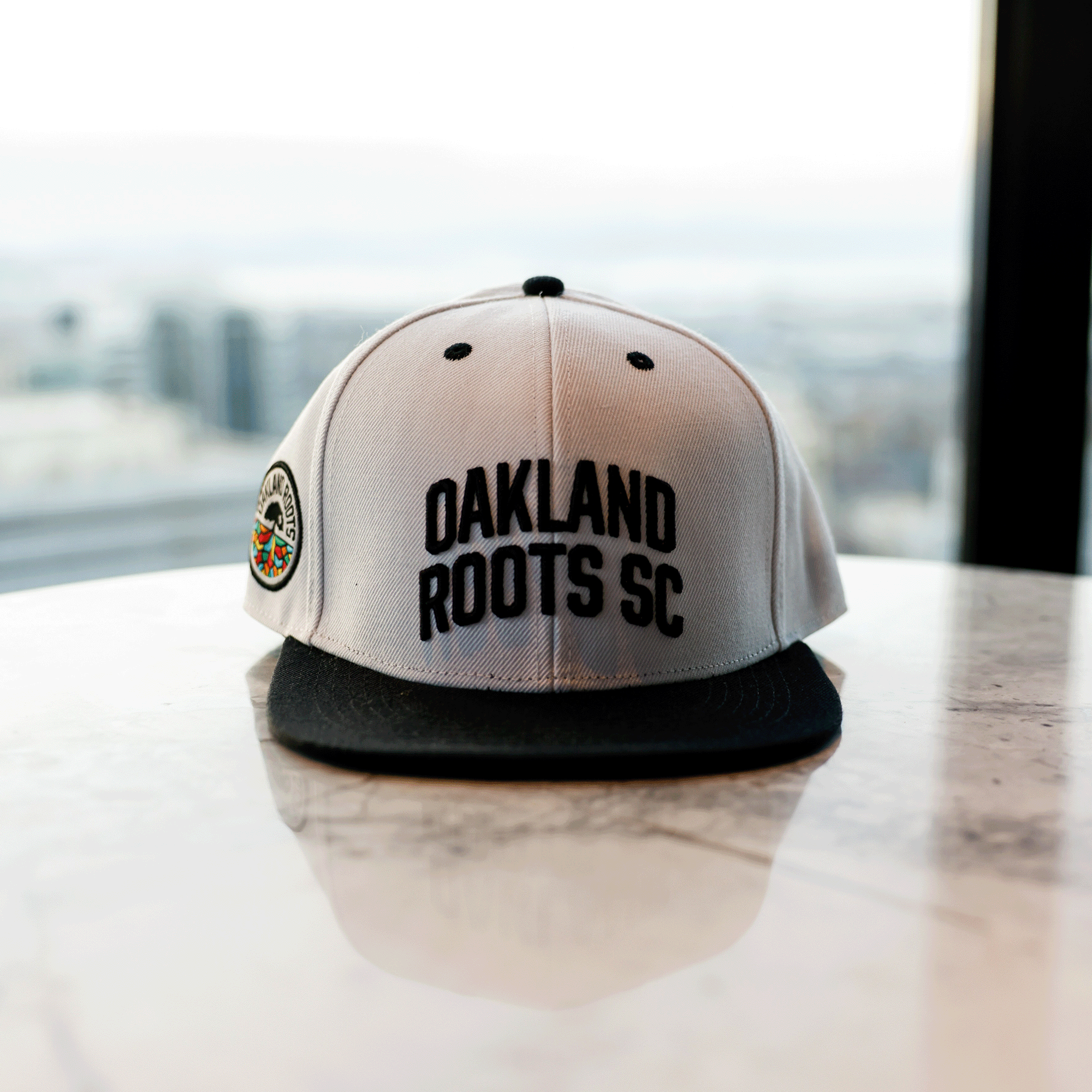 An Oakland Roots SC Arched Text Snapback in white and black, featuring "OAKLAND ROOTS SC" embroidered in black on the front. The cap also showcases a round, multicolored logo on the side. It is placed on a reflective marble surface with a blurred cityscape visible through large windows in the background.