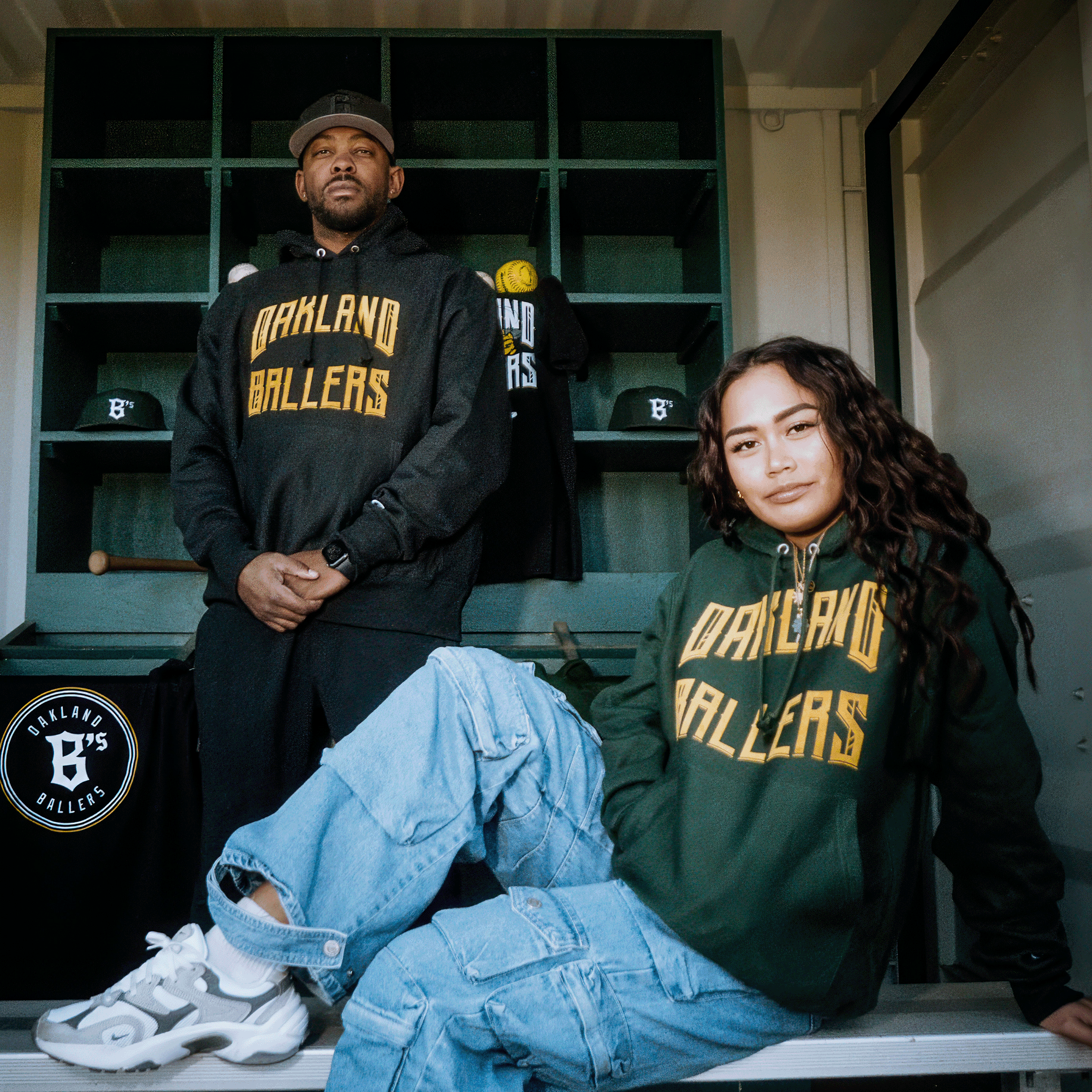 Two people in Champion X Oakland Ballers Wordmark Hoodies stand before a shelf of hats and shirts. The black-clad left figure clasps their hands, while the green-clad right sits with a bent leg. They're proudly showcasing Oakland Ballers' new pro baseball team, with its logo displayed on the wall.