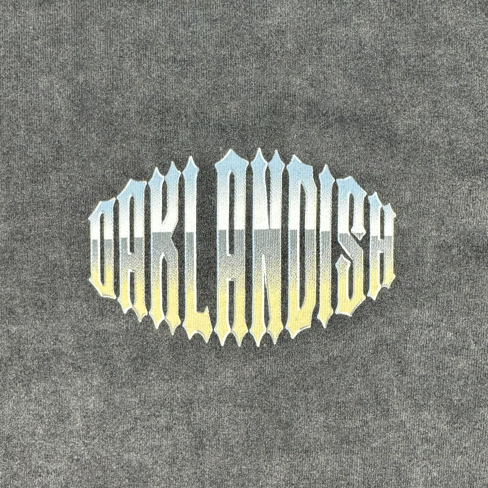 The Chopper Heavy LS Tee features bold "OAKLANDISH" text on a textured gray, vintage black long sleeve shirt. The letters have a gradient from blue to yellow, emulating the sky. Made with 100% cotton for comfort and durability.