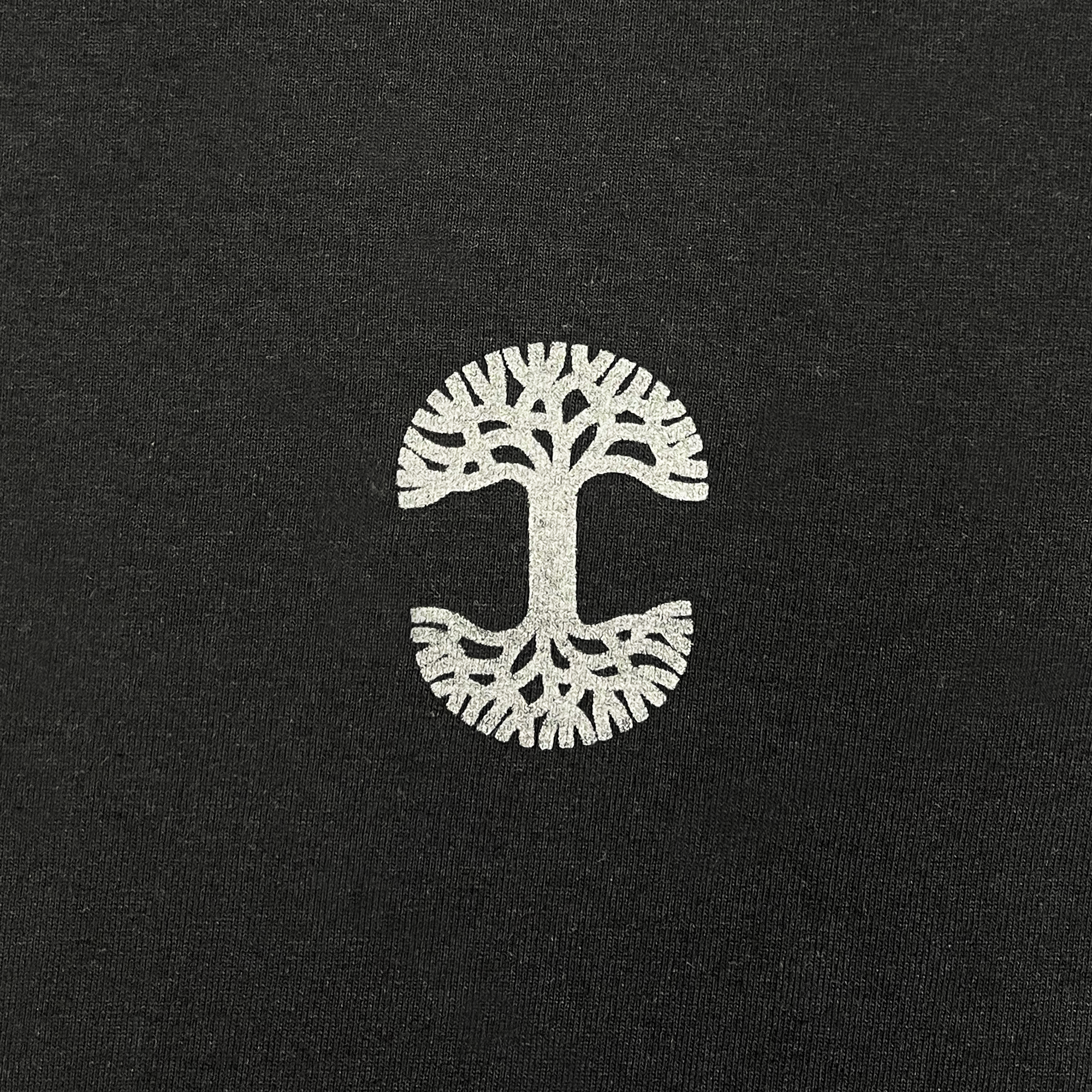 The Cipher LS Tee by Oaklandish showcases a black background with a white tree logo on a classic fit long sleeve t-shirt. The branches of the tree spread at the top, while its roots mirror them at the bottom. The intricate design is printed in reflective ink, forming a horizontally elongated "T" shape at the center on 100% cotton fabric.