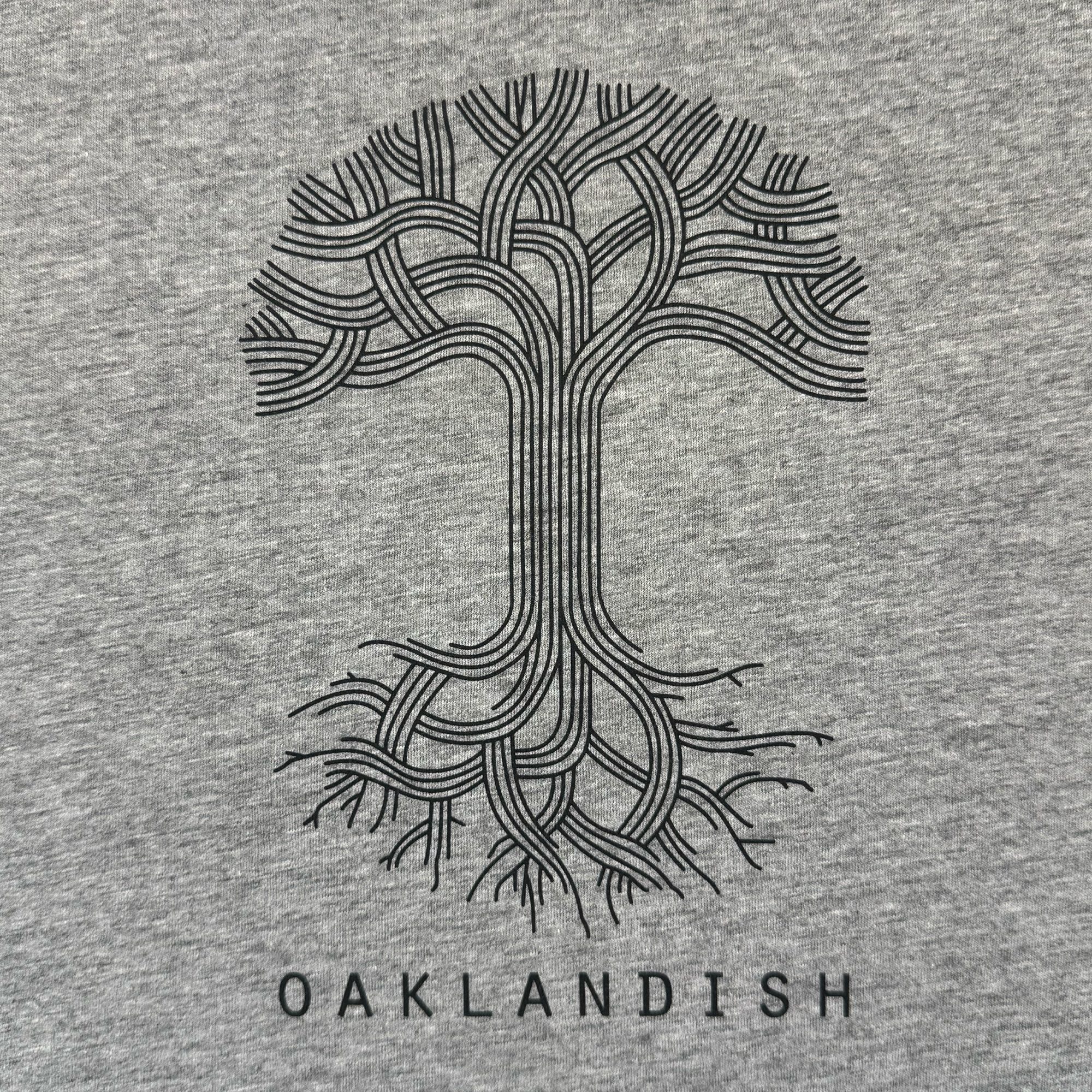 The Women's Oaklandish Logo Tee by Oaklandish features a stylized black tree with intertwining branches and roots centered on a classic fit grey 100% cotton shirt, with "OAKLANDISH" in bold capitals beneath the design.