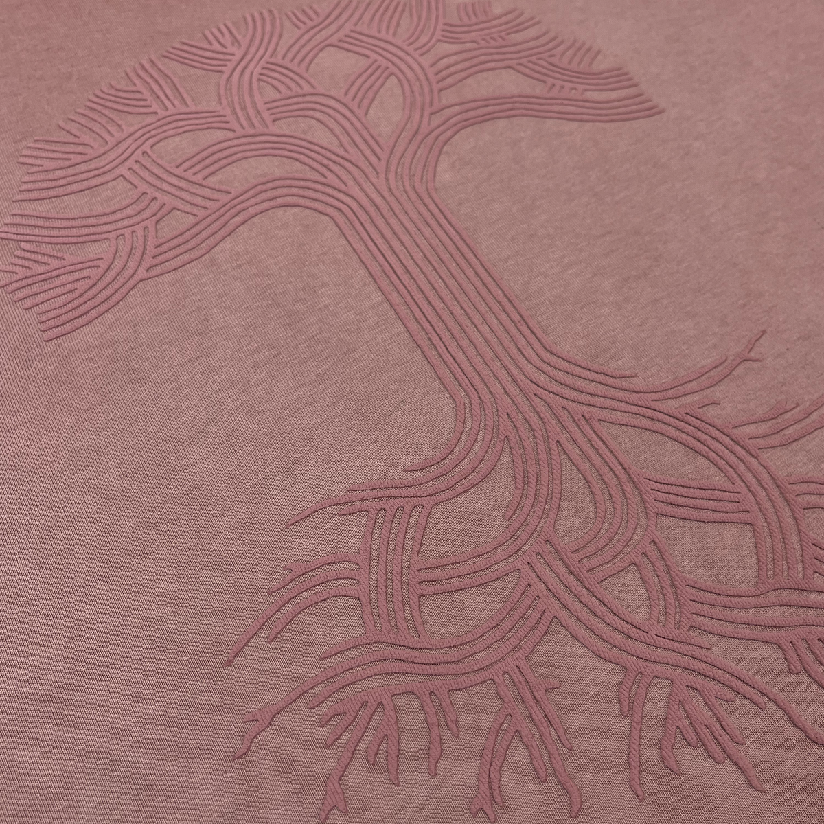 Close-up of the Oaklandish Classic Logo Tee by Oaklandish, featuring a textured design on heavyweight cotton fabric. The iconic tree logo, with its intricate, winding branches and roots formed by parallel raised lines, stands out in a slightly darker shade against the dusty pink fabric. This subtle contrast adds visual depth—making it an ideal choice for a classic fit men’s t-shirt.