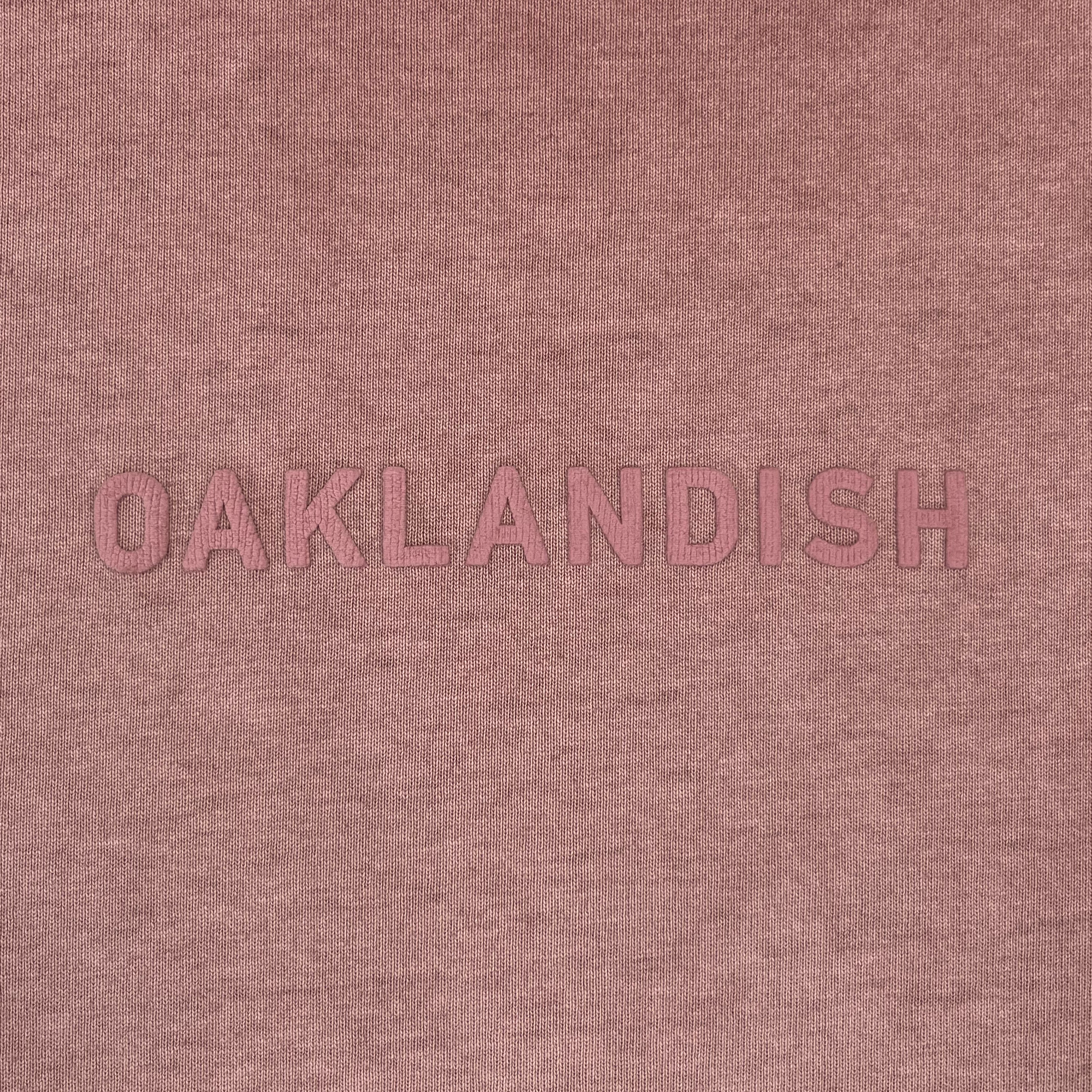 The Oaklandish Classic Logo Tee by Oaklandish features bold, uppercase “OAKLANDISH” lettering in pink, centered on a soft, textured dusty rose or mauve heavyweight cotton background. The minimalist, monochromatic design offers subtle contrast and comes in a classic fit men’s sizing.