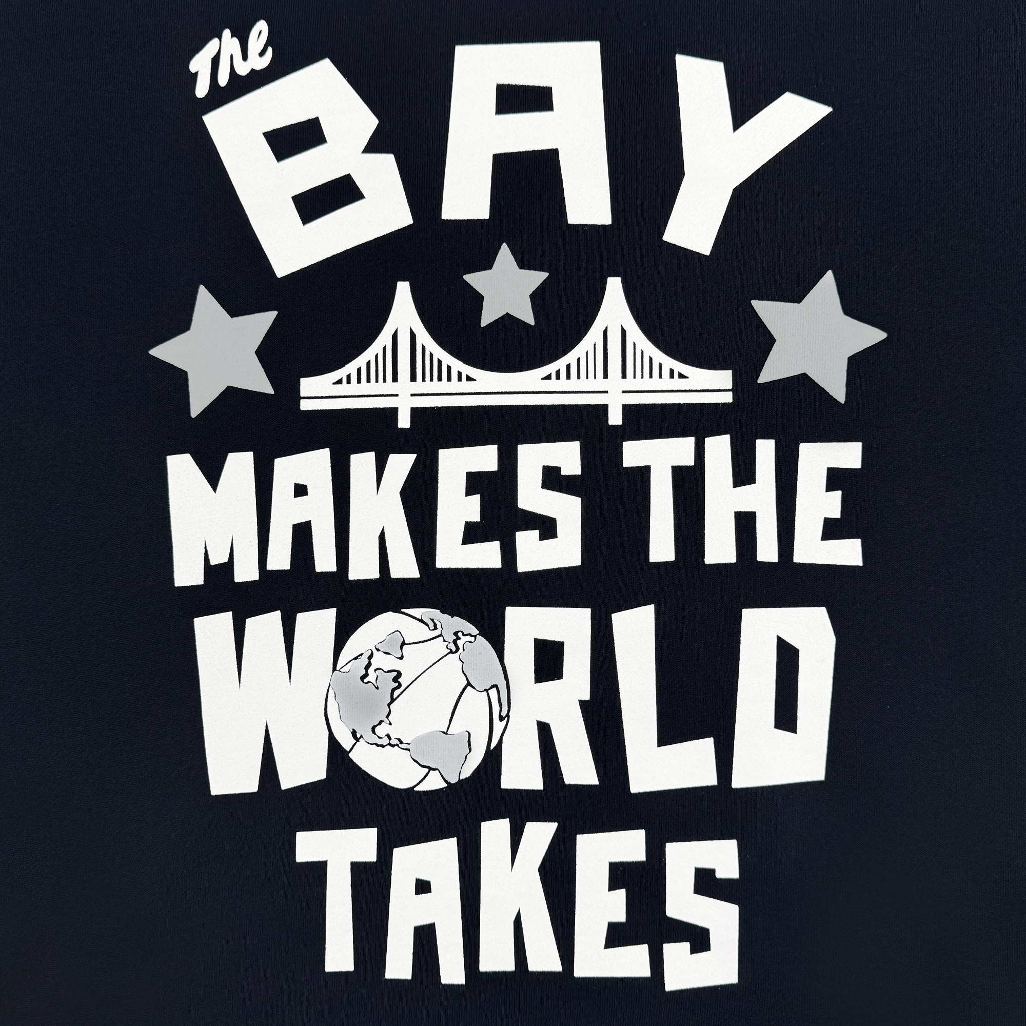 The Bay Bridge Hoodie by DOC from Oaklandish features the text "The Bay Makes The World Takes" in stylized letters. Above, a bridge graphic flanked by stars honors Bay Area culture, with a globe illustration below. Designed by Dustin O. Canalin on a black background with white graphics and text.