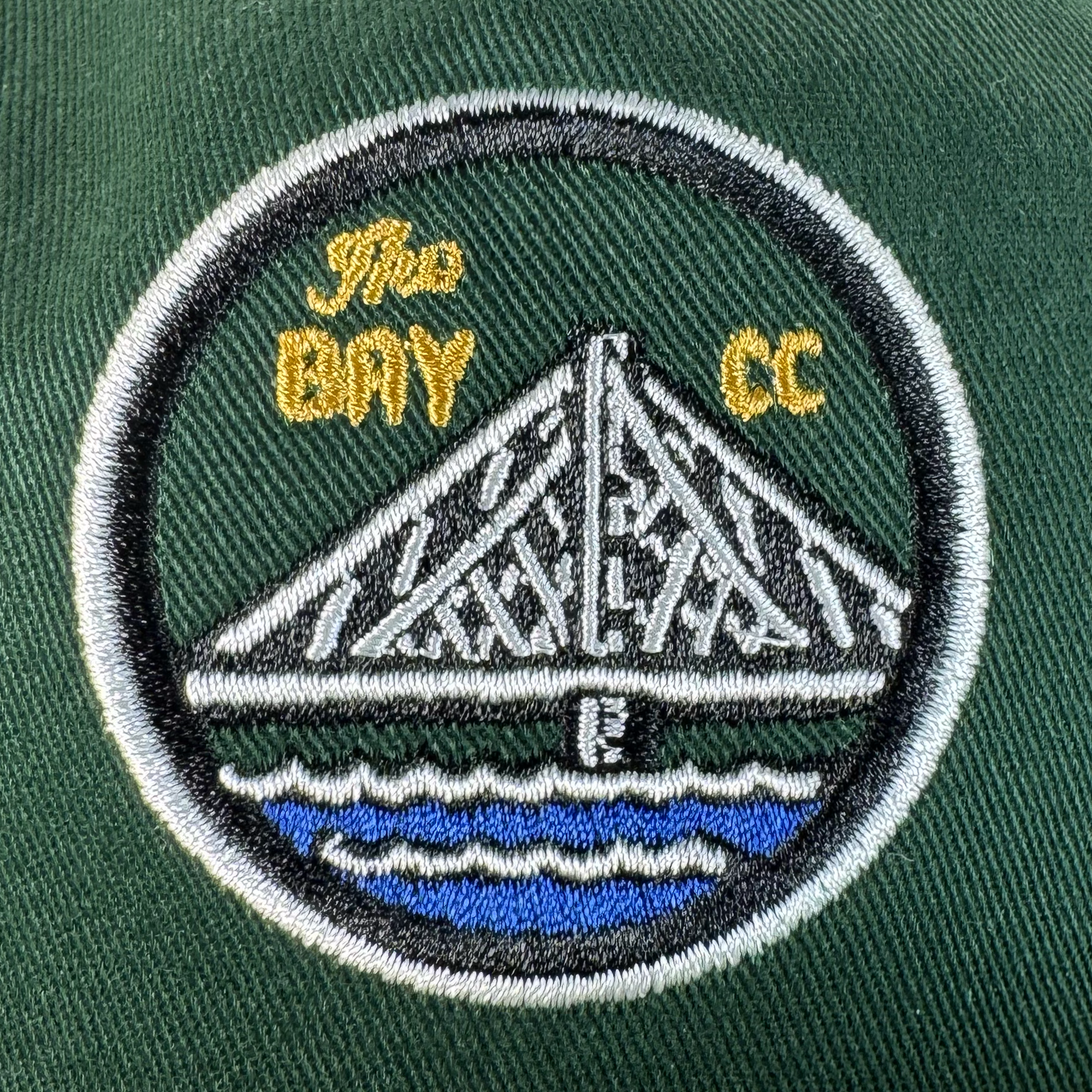 The Bay Area 5 Panel By DOC from Oaklandish features an embroidered white bridge over blue water on dark green fabric, bordered in black and white with "The BAY CC" in yellow—perfect for your favorite 5-panel hat.