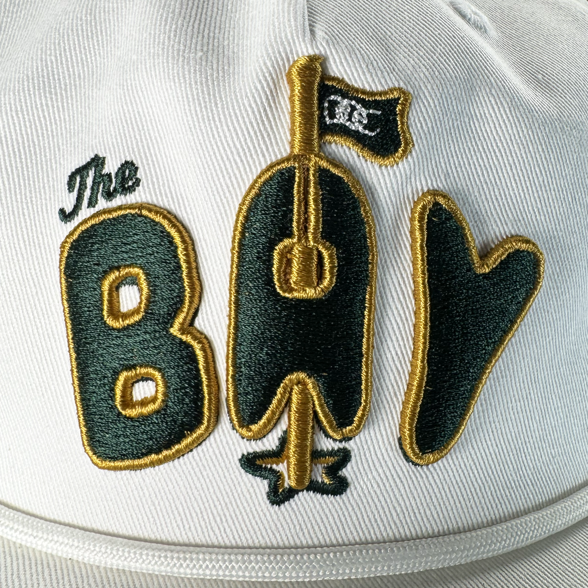 Close-up of the Oaklandish Bay Area 5 Panel By DOC hat: a white, unstructured strapback with detailed stitching, featuring bold dark green and gold "The Bay" embroidery—where the "A" forms a guitar and the "Y" resembles a heart.