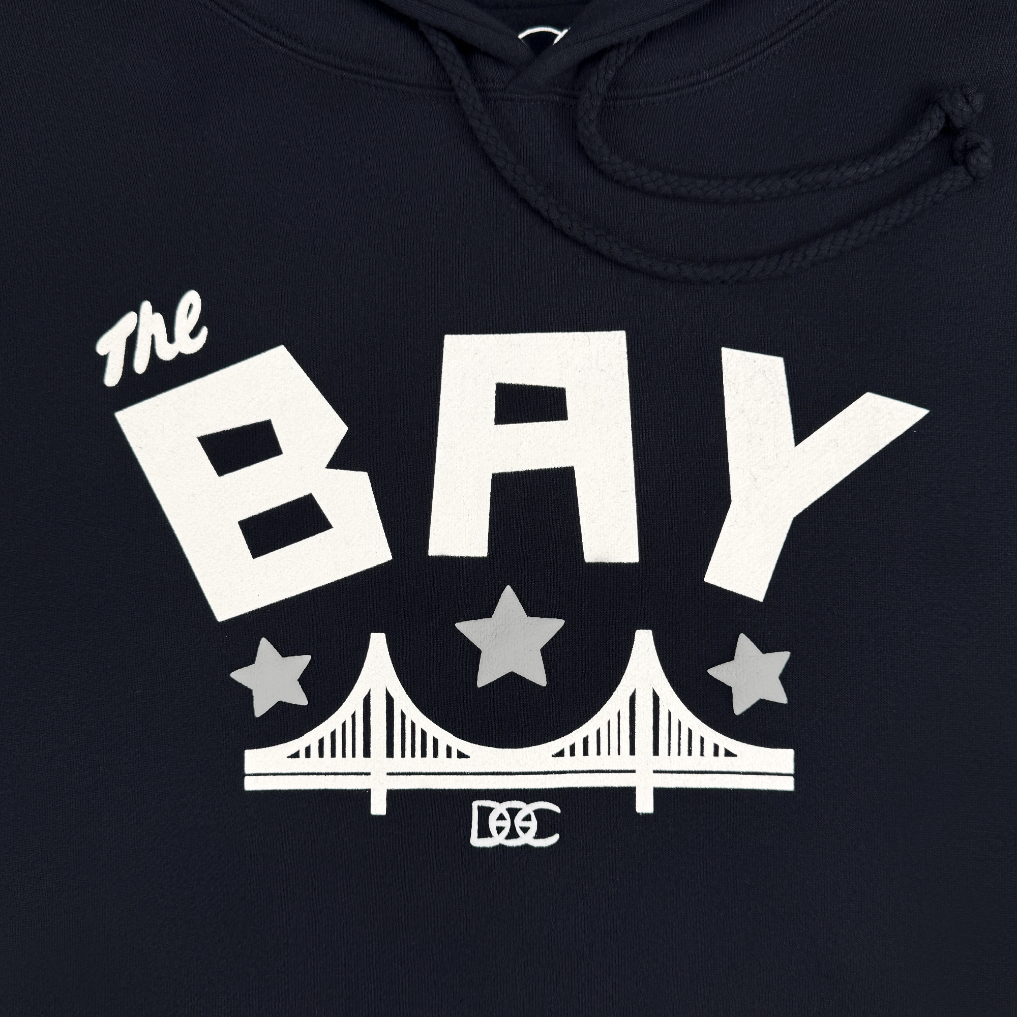 The Bay Bridge Hoodie by DOC from Oaklandish features a bold white graphic design with "The" in cursive and "BAY" in block letters. Two gray stars flank a bridge illustration by Dustin O. Canalin, with a smaller star above and the "DC" logo below, inspired by Bay Area vibes.