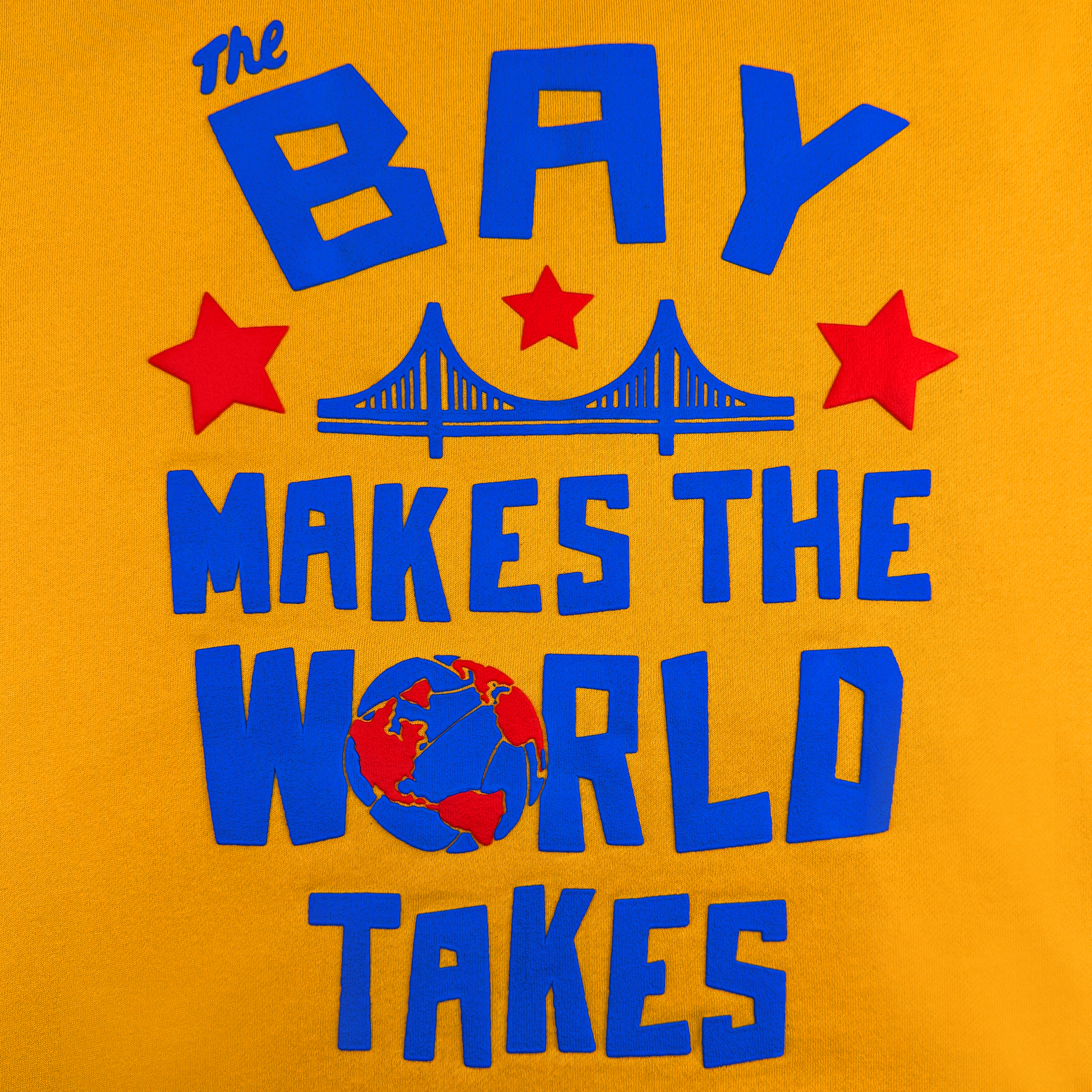 Text on a bright yellow background reads, "The Bay Makes The World Takes" in bold blue letters, showcasing the DOC Bay Bridge Hoodie by Oaklandish. Three red stars and a stylized blue bridge symbolize the Bay Area's cultural influence, with a red and blue globe reminiscent of an art project below.