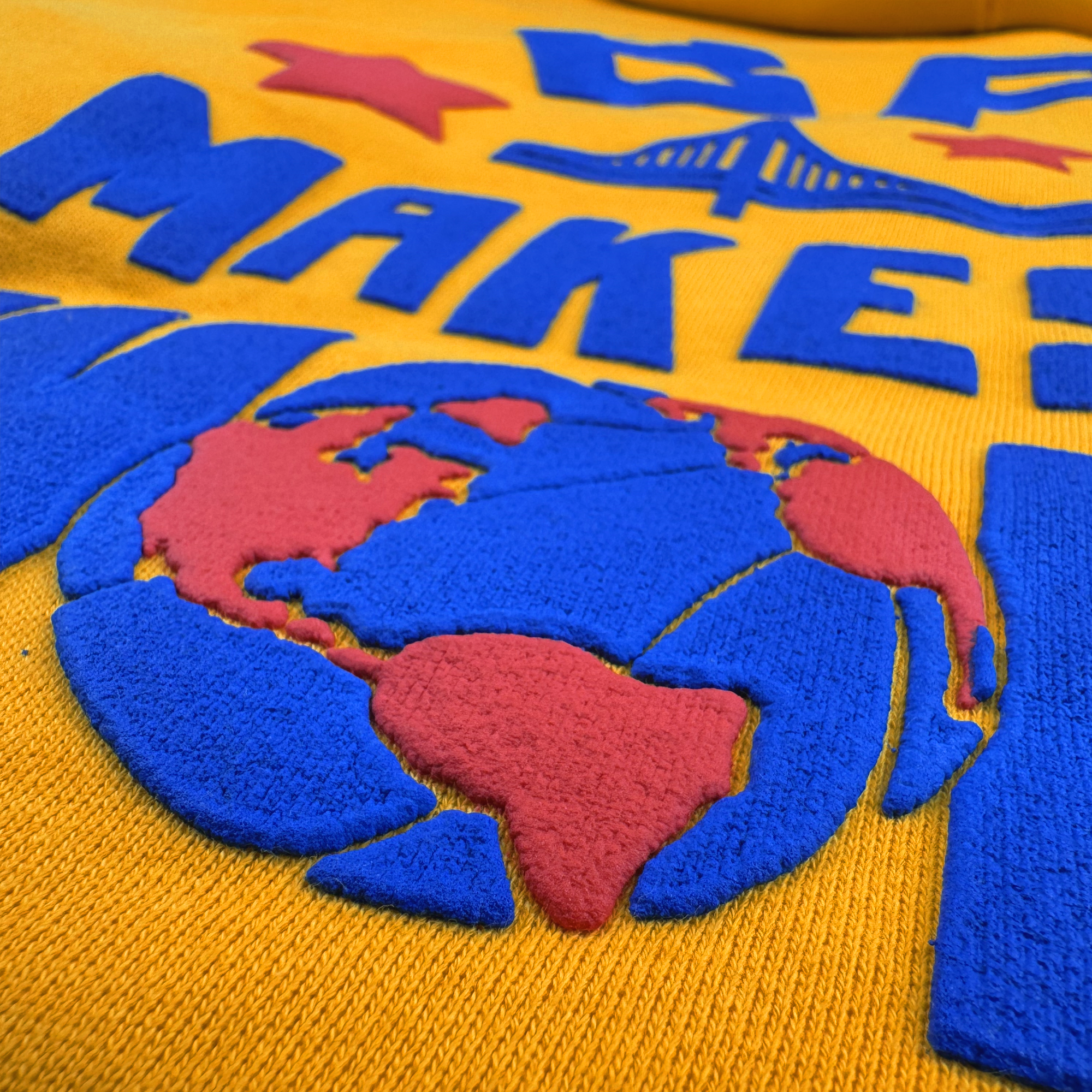 The Bay Bridge Hoodie by DOC from Oaklandish features a textured yellow fabric with a blue and red globe, reflecting cultural influence. Above it, "MAKE" is spelled in blue letters. The design includes red stars and a stylized bridge inspired by the Bay Area art project scene.