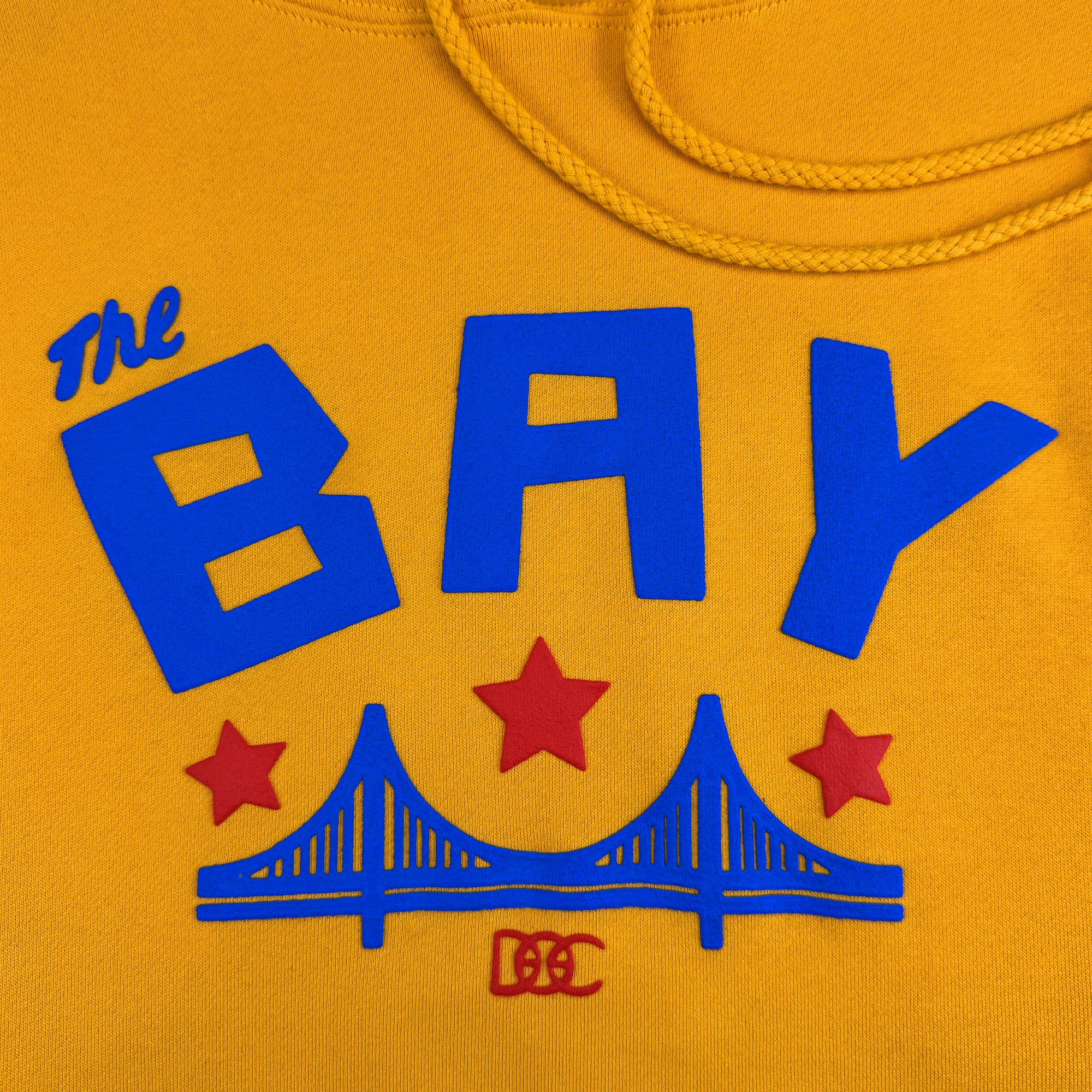 The Bay Bridge Hoodie by DOC from Oaklandish features blue "The Bay" text, a bridge graphic with three red stars, and the initials "DC" on a yellow background. A drawstring completes this vibrant tribute to the Bay Area's cultural influence.