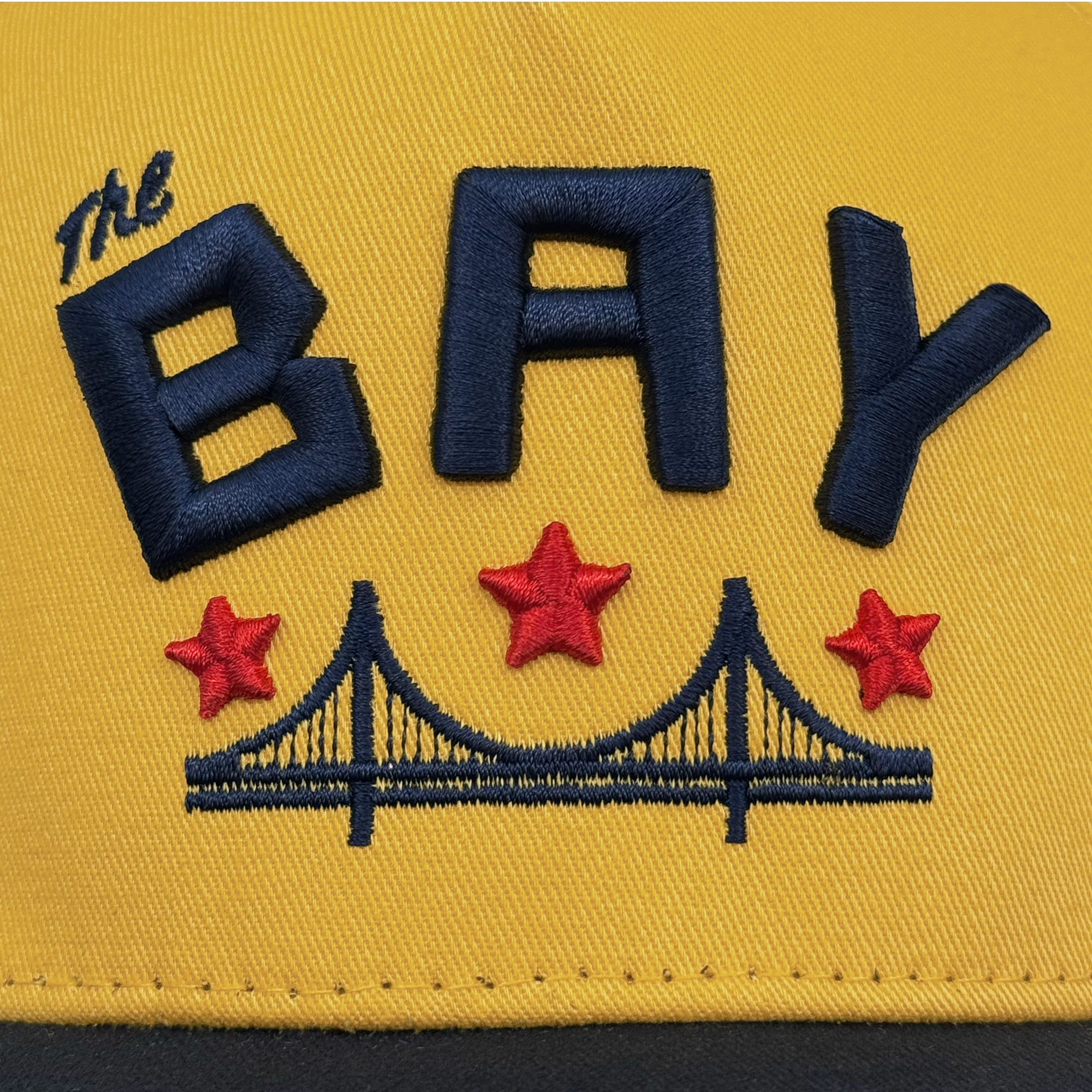 The Bay Bridge Snapback by DOC from Oaklandish is a masterpiece of art and design with "The BAY" in bold black letters embroidered on yellow fabric, featuring a stylized Bay Area bridge and three red stars.