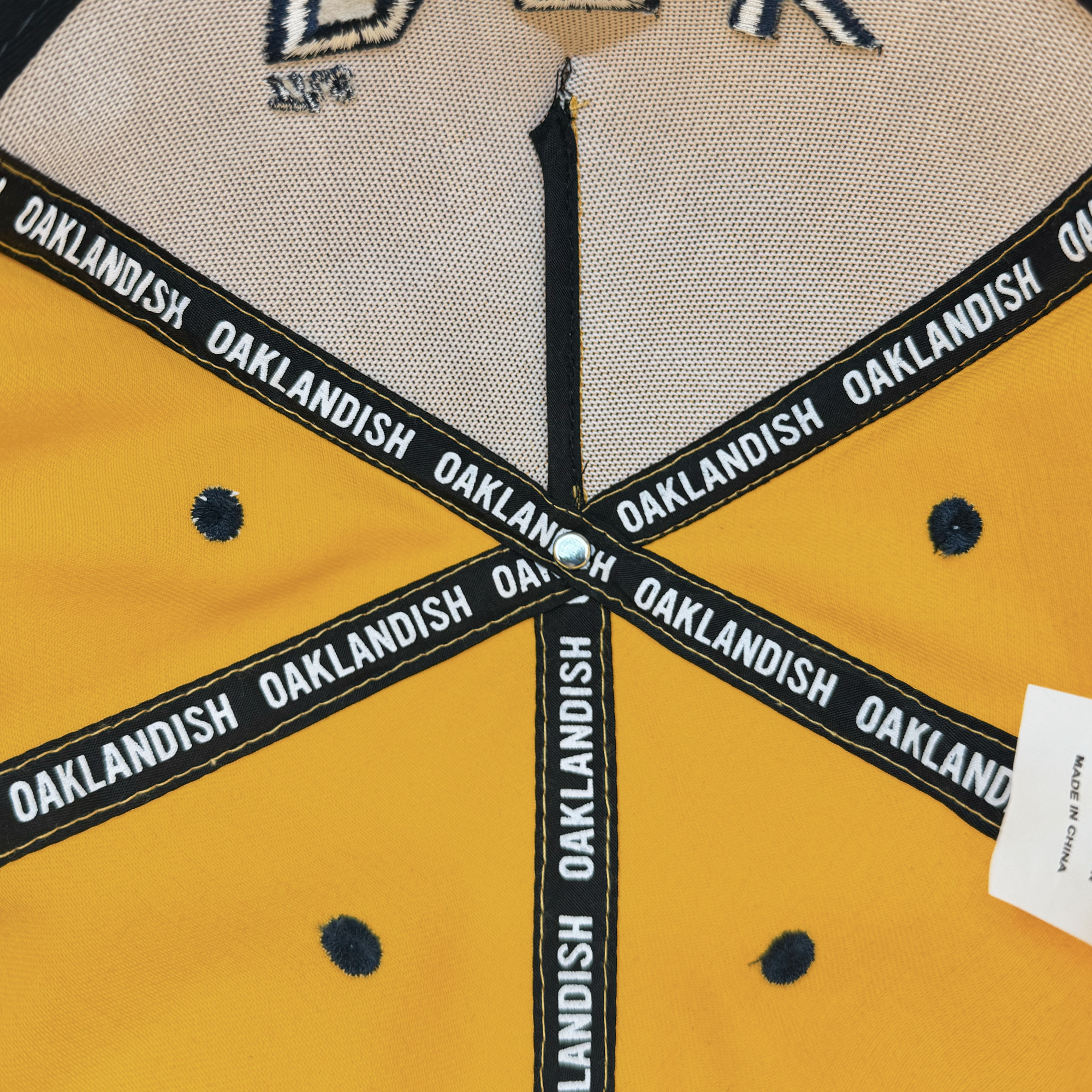 Close-up of a yellow Bay Bridge Snapback by DOC with black polka dots, intersecting seams bearing "Oaklandish" in white text, and a white "MADE IN CHINA" tag at the bottom right.