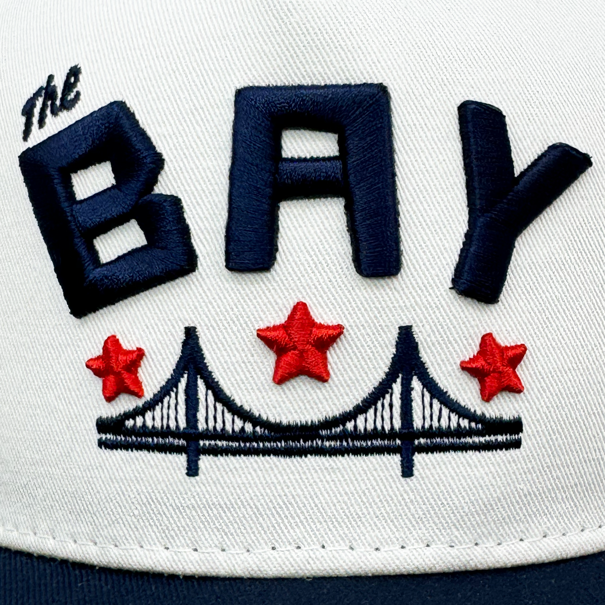 The Bay Bridge Snapback by DOC, crafted by Oaklandish, showcases "The BAY" in bold blue embroidery with three red stars above a blue and white suspension bridge outline reminiscent of the Golden Gate, capturing Bay Area culture perfectly on its front.