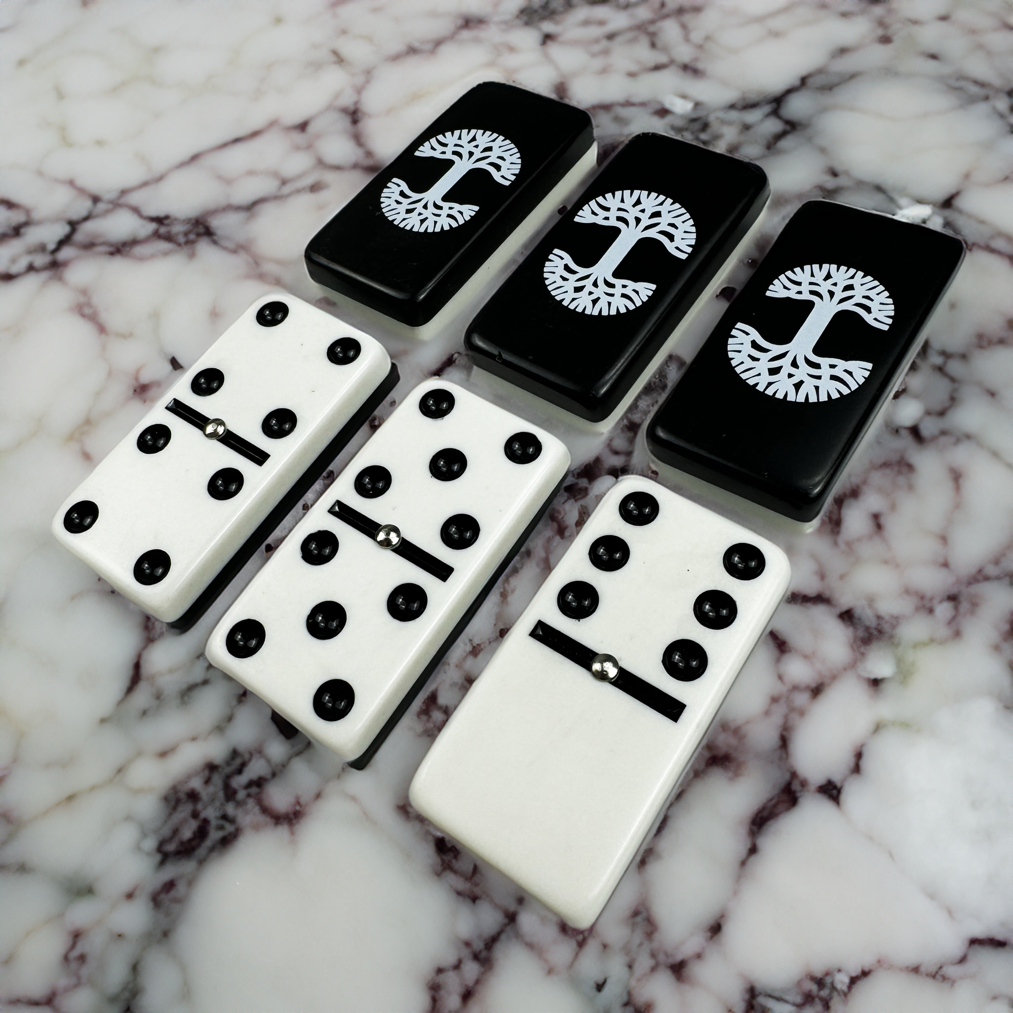 Oaklandish Dominoes Treasure Chest on a marble surface. The top row displays black tiles adorned with white Oaklandish tree designs, while the bottom row showcases white tiles with black dots in various arrangements, including two double-threes and one double-four.