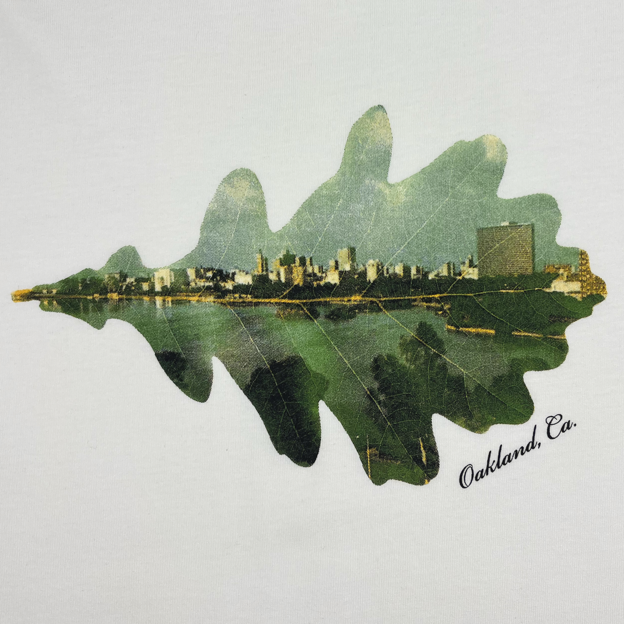 Introducing the Double Exposure Tee by Oaklandish: This design features an image of a leaf with an artistic overlay of the Oakland, California cityscape. The city's skyline, reflecting on water, is beautifully displayed within the outline of the leaf. "Oakland, Ca." is elegantly written in cursive at the bottom right of the leaf. Printed on a 100% cotton classic fit t-shirt available at Natural Shop. The background is white.