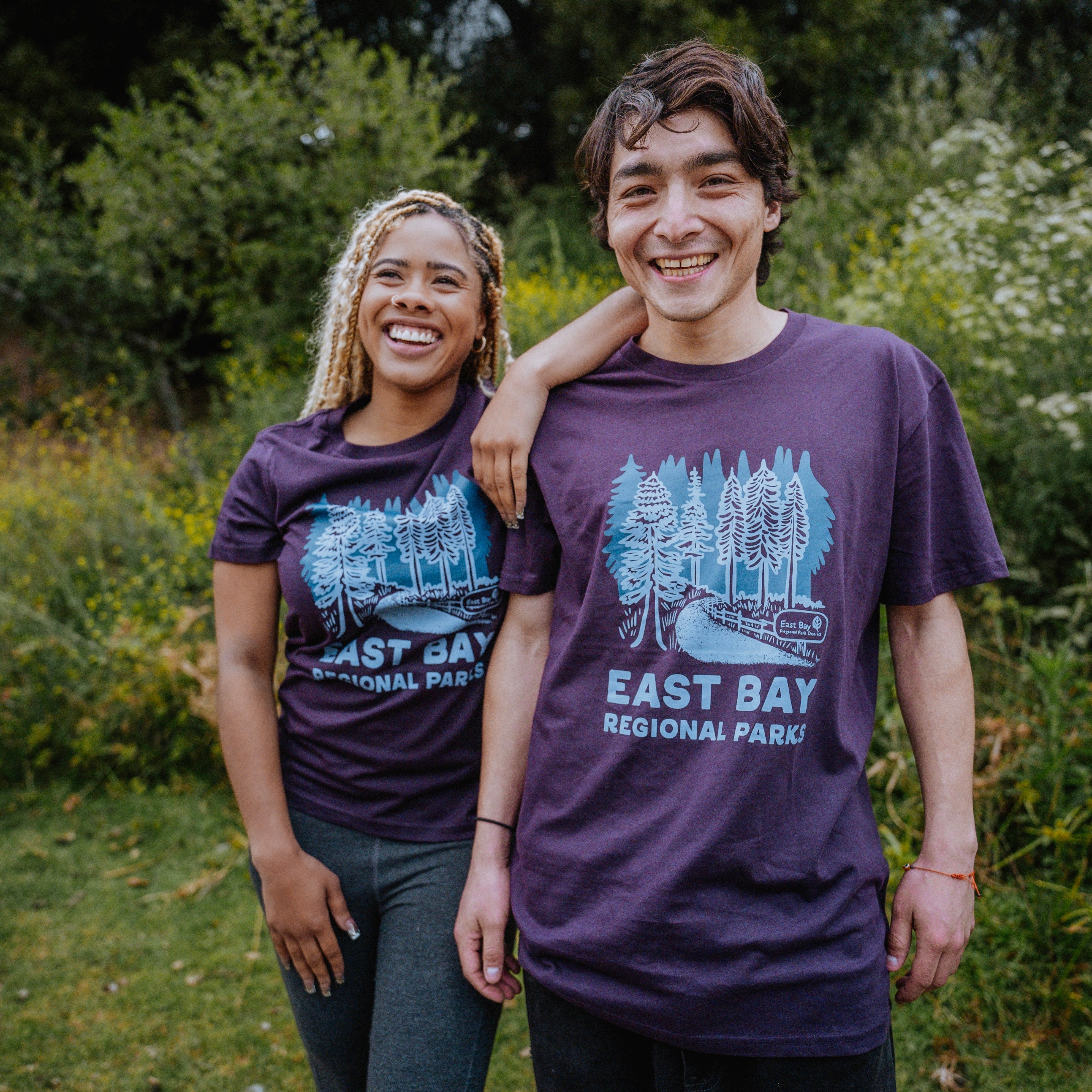 Two people standing outdoors, both wearing purple "Women's East Bay Regional Parks Tee" from Oaklandish. The person on the left has curly blond hair and is leaning on the shoulder of the person on the right, who has short dark hair. They are both smiling, with greenery and trees in the background.