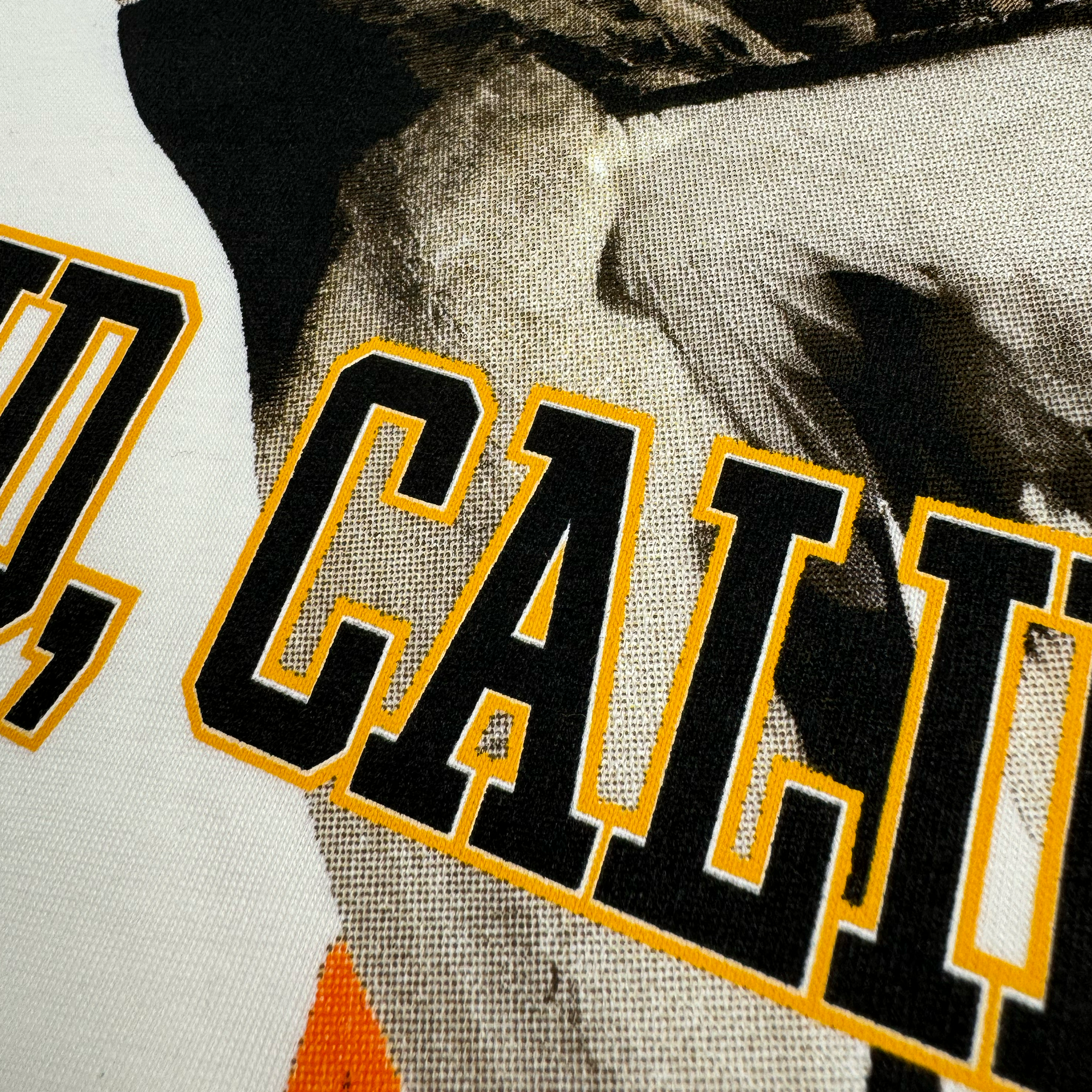 Close-up of a textured 100% cotton fabric print featuring the words "OLD CALI" in bold, black letters with a yellow outline. Overlaid on this text is a black and white, grainy image, possibly of a person. The fabric exudes a vintage design with visible weave detail, suggesting that it is likely part of the Oaklandish Duck School Tee.