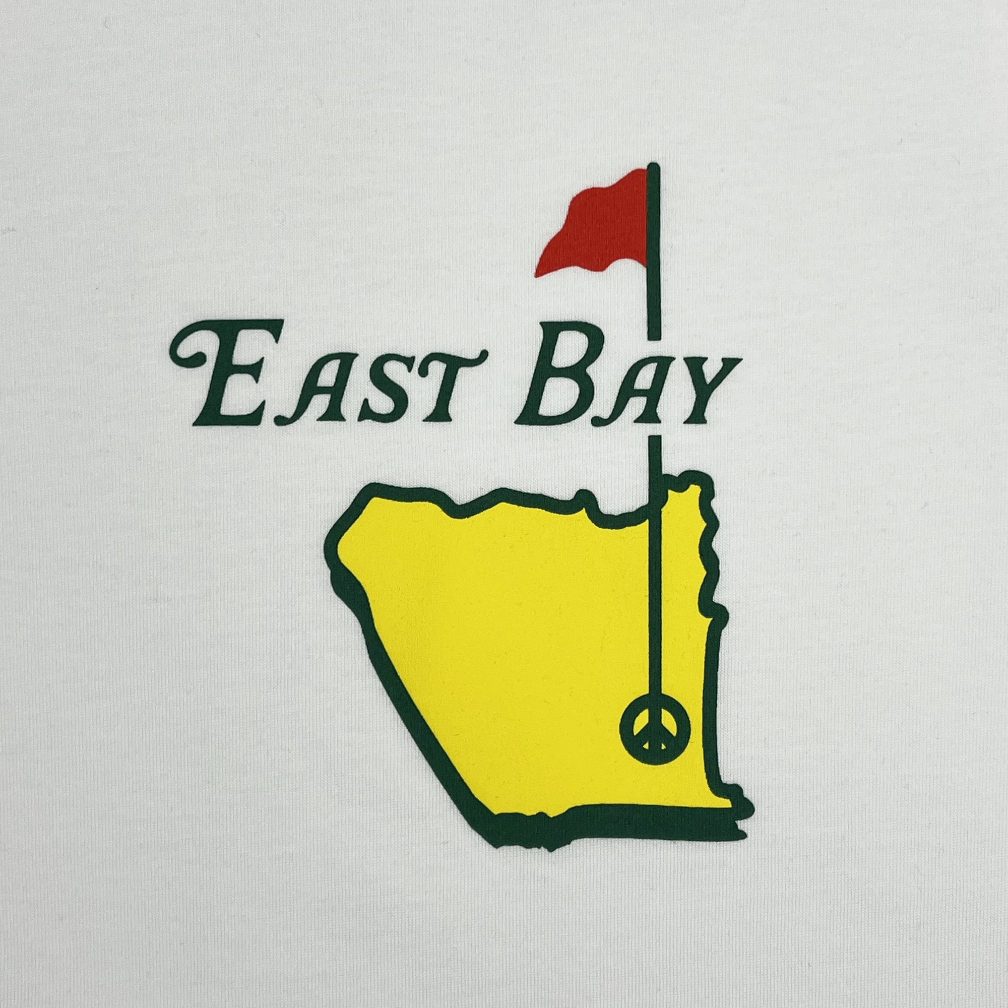 The East Bay Golf Tee by Oaklandish showcases a graphic with "East Bay" in dark green text, accompanied by a yellow map outline beneath it. A red flag appears on the upper right side of the map, featuring a golf hole marker shaped like a peace symbol. The design is set against a white background and crafted from 100% cotton for a classic fit t-shirt.