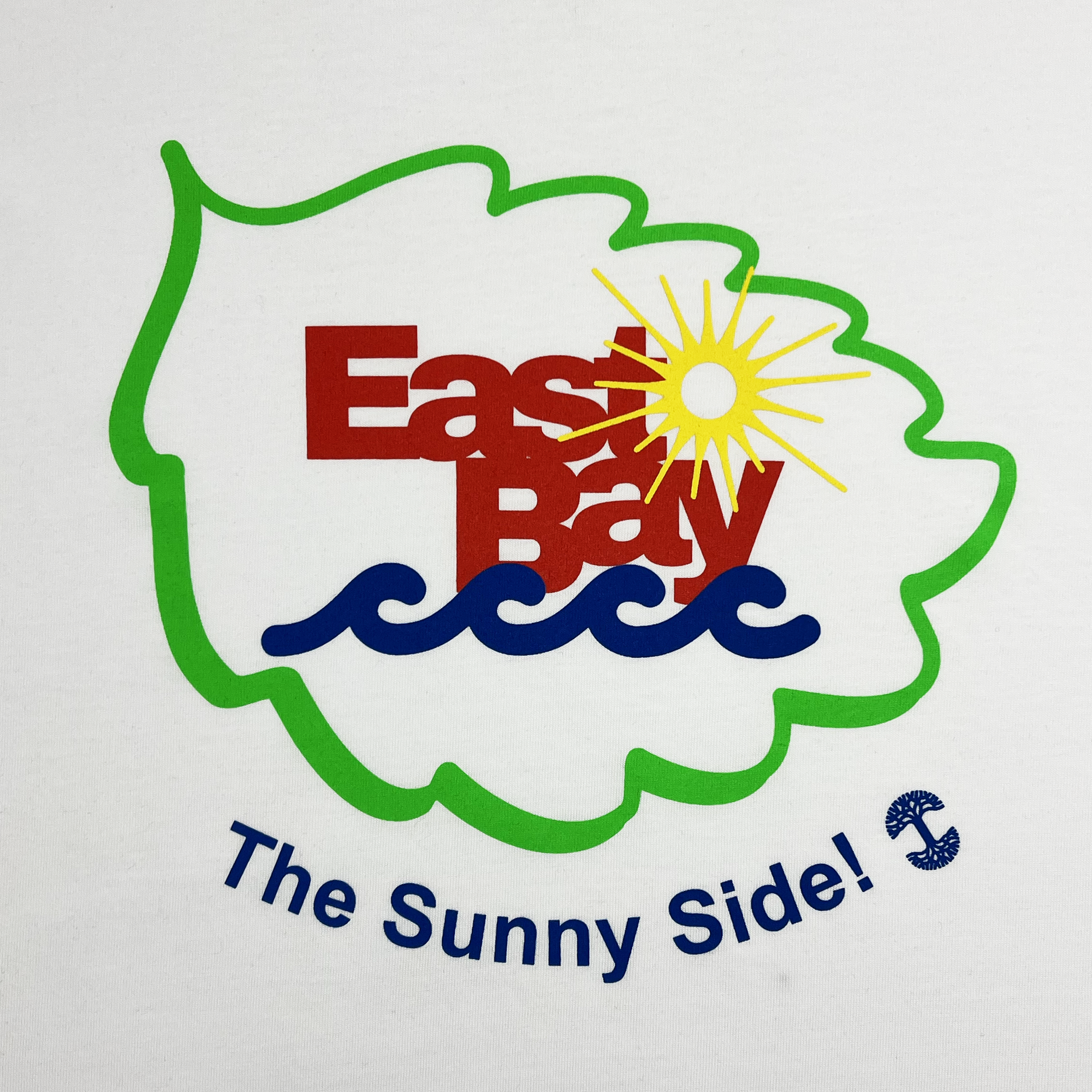 The East Bay Sun Tee by Oaklandish features a green leaf outline with the words "East Bay" in bold red letters at the center. The design incorporates a yellow sun and blue waves, with the slogan "The Sunny Side!" written in blue text below the leaf and a small anchor symbol replacing the letter “i” in “Side!”. This classic fit t-shirt is part of our men's collection.