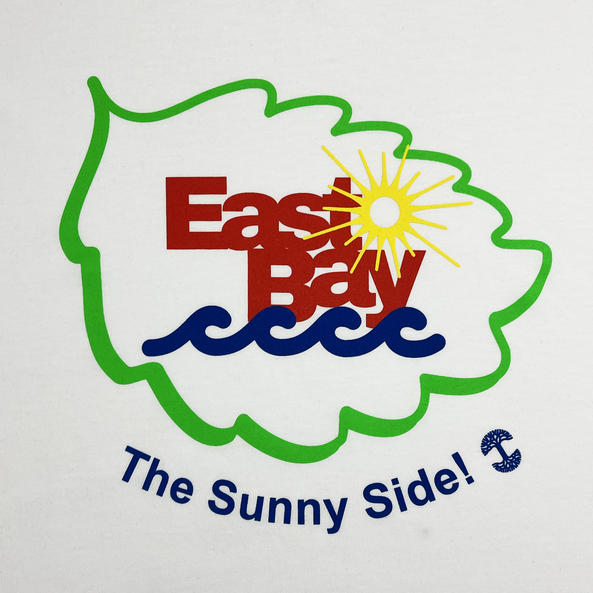 Introducing the East Bay Sun Tee by Oaklandish: This men's 100% cotton t-shirt features a green leaf outline with "East Bay" in bold red letters. Above the design, a bright yellow sun with rays illuminates blue wave patterns, while beneath the leaf, "The Sunny Side!" is accompanied by a small blue anchor. The entire design sits on a pristine white background.