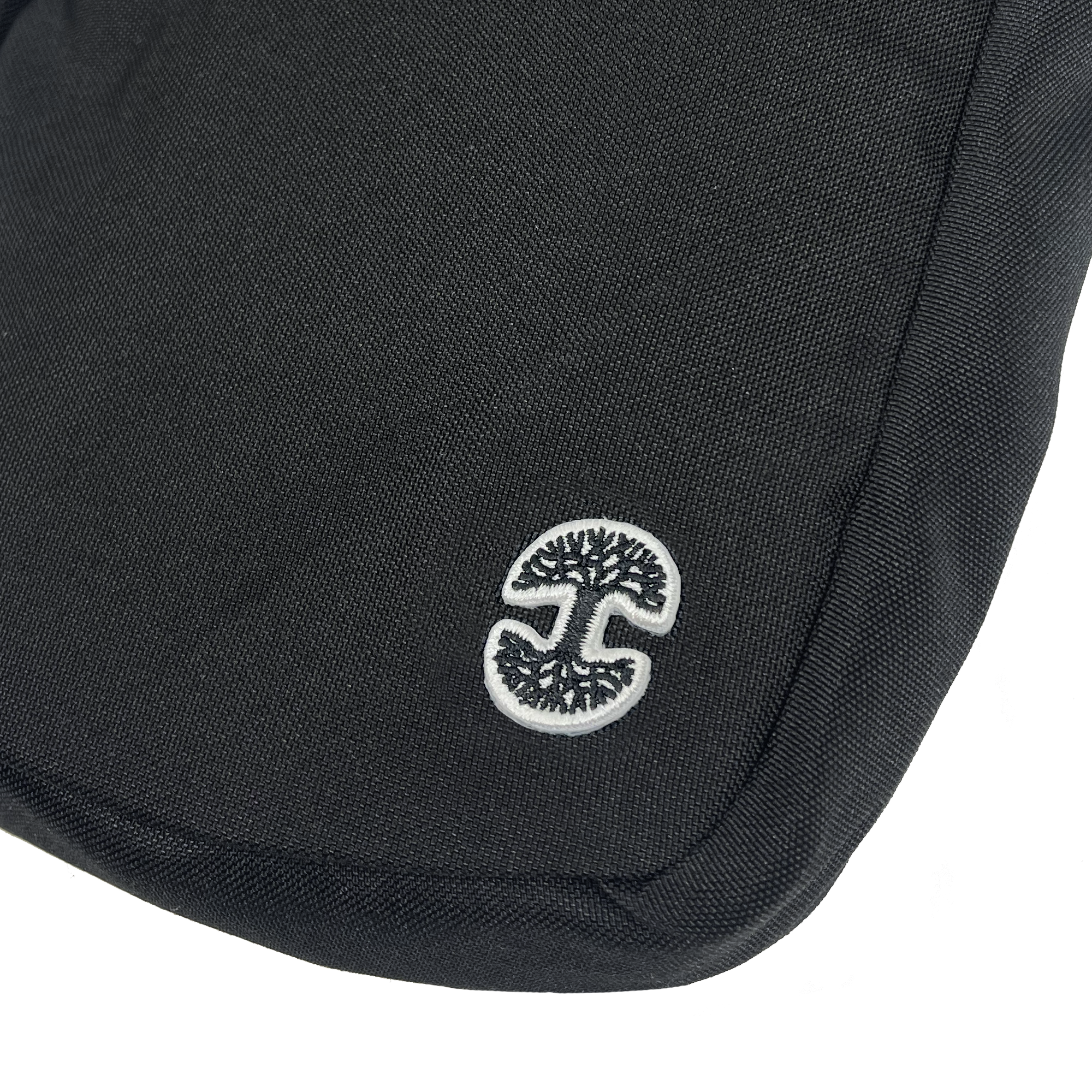 A close-up of the Oaklandish Flight Bag reveals its black textured weave fabric, adorned with a small, white embroidered Oaklandish tree logo. The logo, a stylized tree encircled within a white border, is positioned on the bottom left corner of the bag.