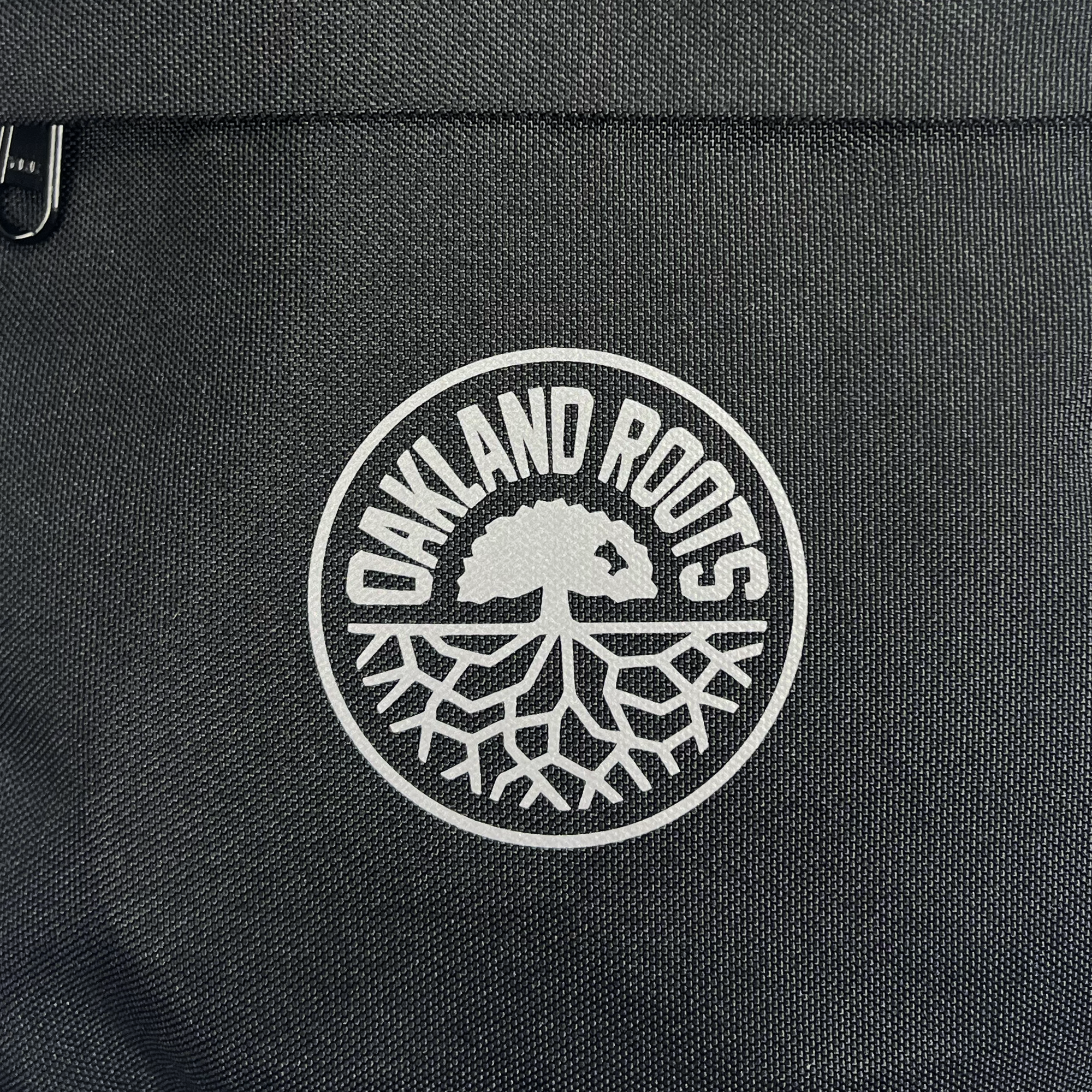 The Oakland Roots SC Flight Bag features a black fabric surface emblazoned with the Oakland Roots logo, symbolizing a commitment to social good. The white circular design on the bag prominently displays "OAKLAND ROOTS" curving around the top, with a stylized tree and its intricate root system below, representing the sports club's deep community connections.