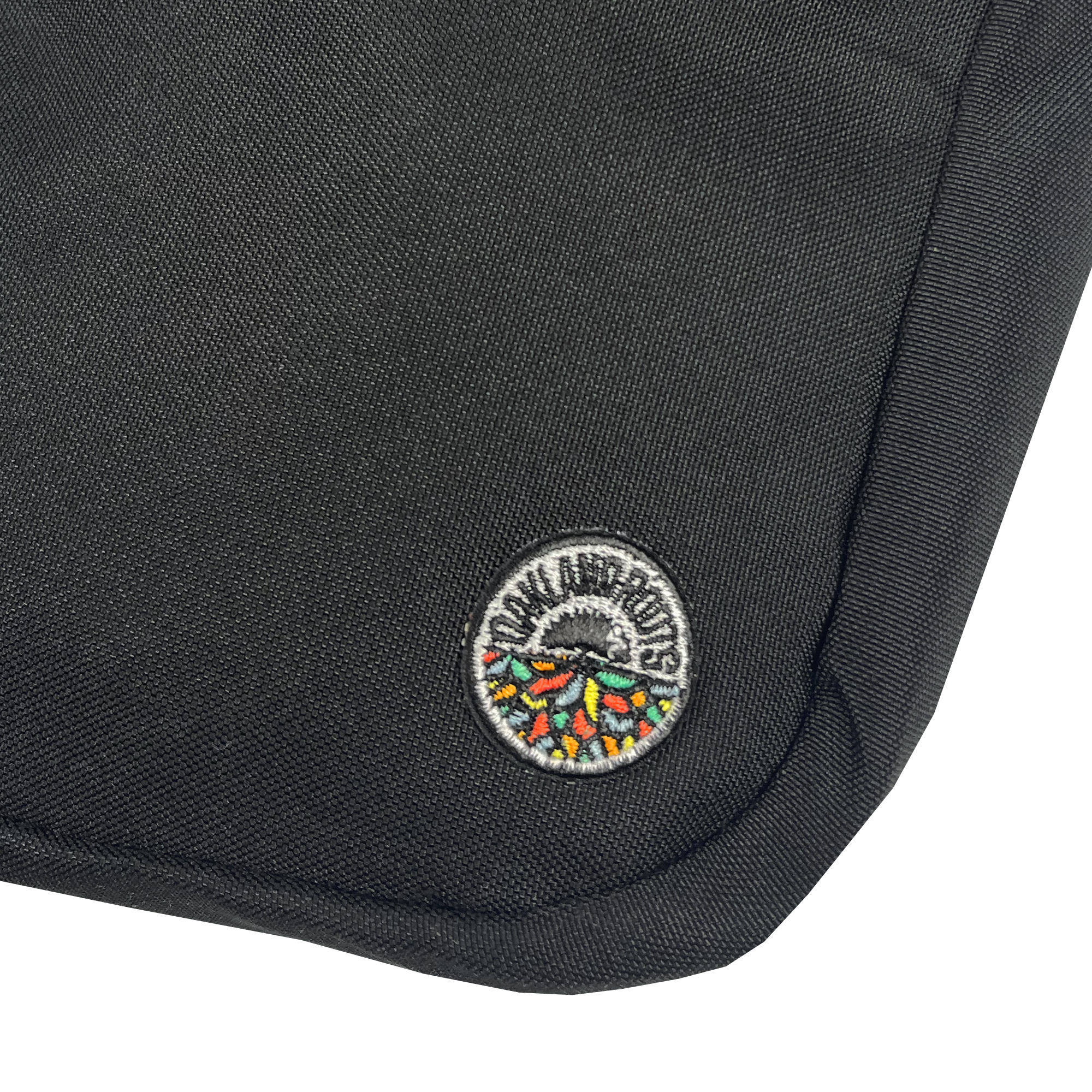 Close-up of the Oakland Roots SC Flight Bag, a black fabric bag featuring a small, round embroidered patch near the bottom corner. The patch showcases a colorful, stylized hedgehog with the text "DON HUSKUTS" around the top. The durable texture and reinforced stitching along the edge make it reminiscent of official gear from an Oakland Roots game.