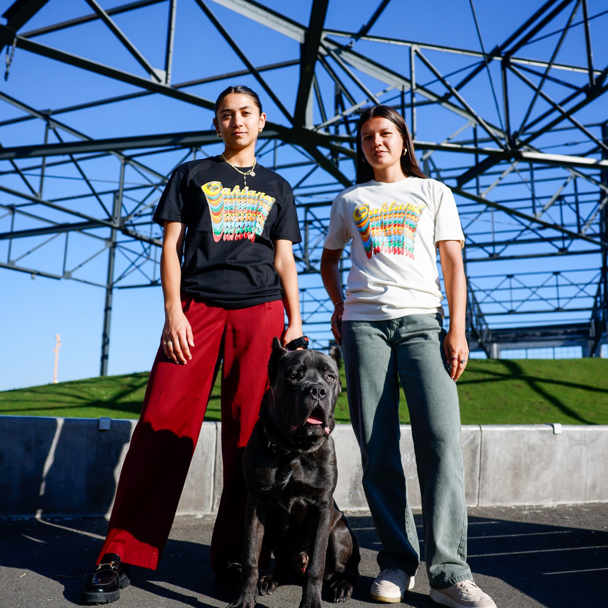 Two people stand outside on a sunny day under a metal structure, with greenery in the background. They both wear Oakland Roots SC Funk Tees and casual pants, showing support for Oakland Roots SC. A large black dog sits between them, looking forward. One person has short hair and wears red pants, while the other has long hair and wears green pants.