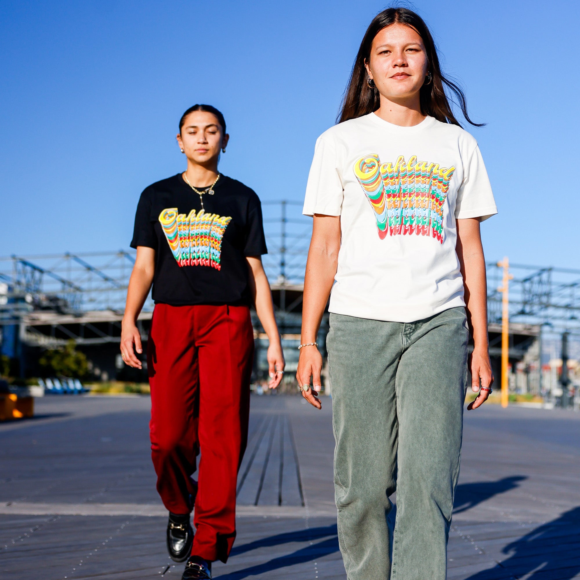 On a sunny day, two individuals walk side by side. The person on the right wears a white shirt adorned with colorful text, light blue-gray pants, and a necklace. The person on the left is sporting an Oakland Roots SC Funk Tee with similar colorful text, red pants, and black shoes. The background features clear skies and a building structure.