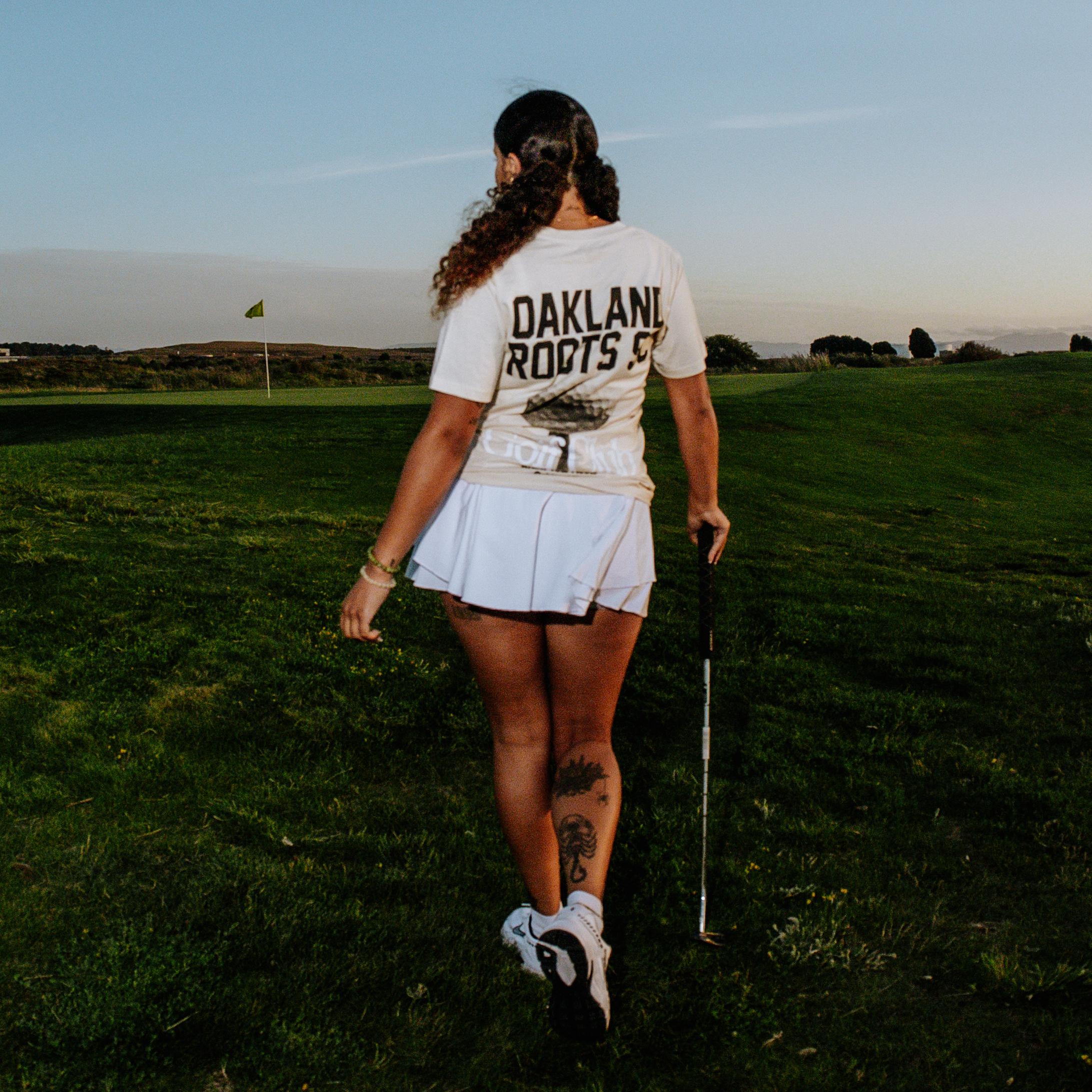 In the golden light of sunset, a woman holding a golf club strides across a serene grassy golf course, embodying the essence of Oakland Roots SC. Clad in a white Golf Club Tee and matching skirt from Oakland Roots SC, her thigh and calf tattoos add a unique touch of personal style. The sky transitions from blue to orange as evening approaches, and in the distance, you can see a golf hole flag waving gently.