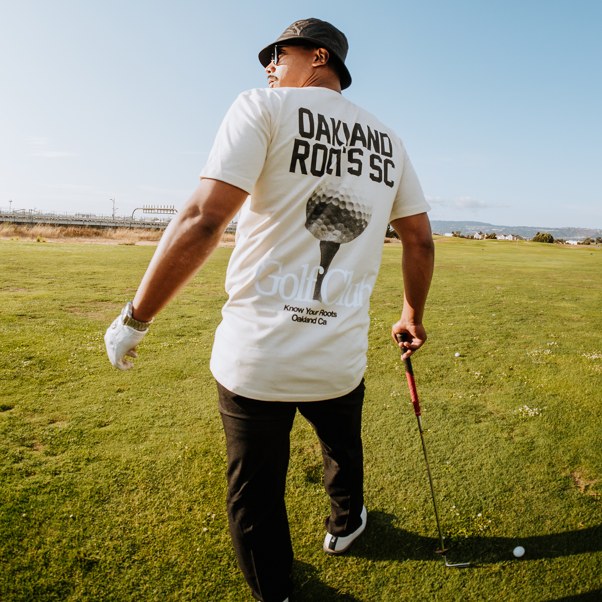 A man wearing a bucket hat and Oakland Roots SC's "Golf Club Tee" featuring "Know Your Roots Oakland CA" on the back stands on a grassy field, holding a golf club. He sports black pants and white gloves, gazing into the distance. The scene is completed by a clear blue sky and low horizon, perfectly capturing the essence of the Oakland Roots Sports Club ethos.