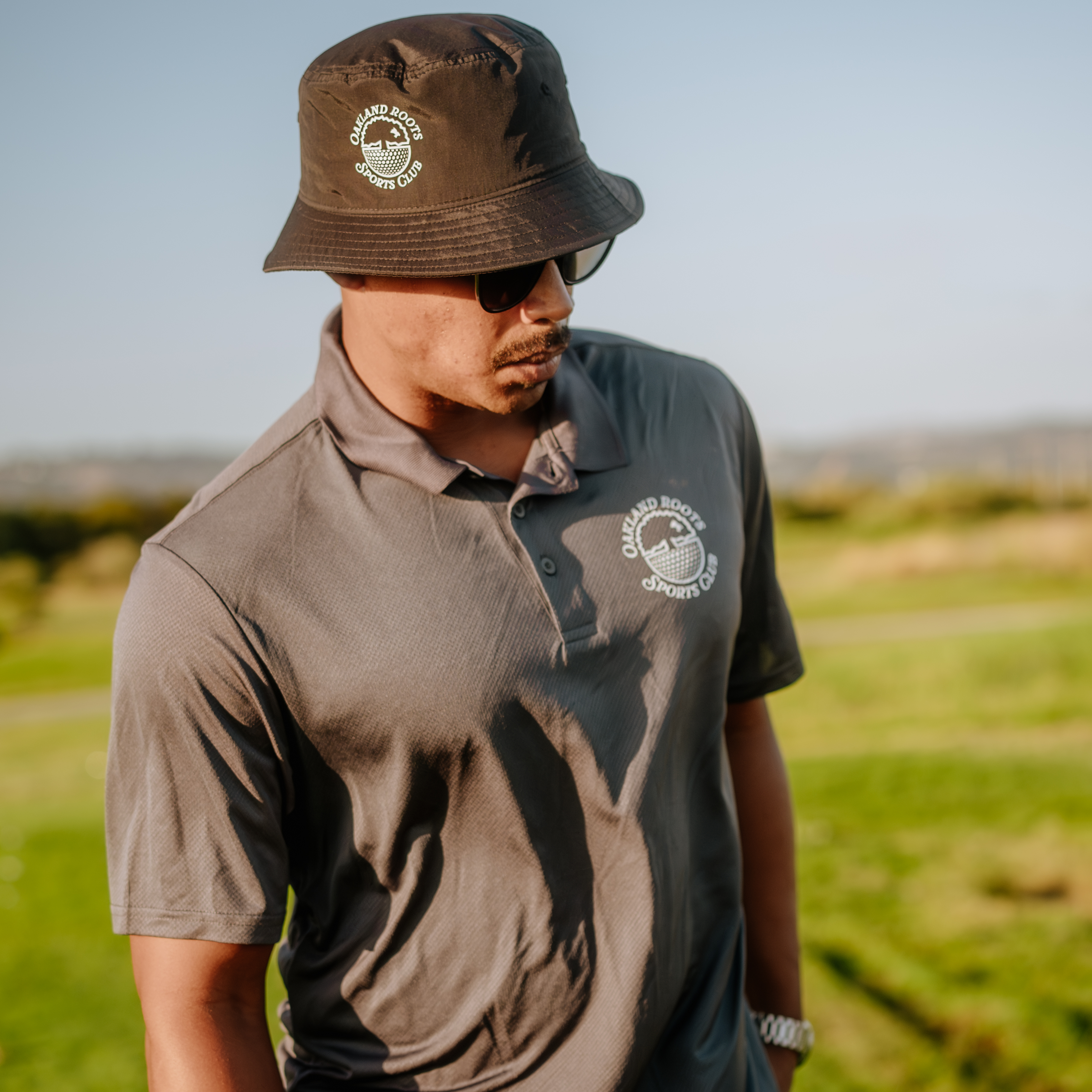A person wearing the Heritage Bucket Hat from Oakland Roots SC, along with sunglasses and a gray polo shirt featuring a circular logo, stands outdoors on a grassy field. Representing the Oakland Roots sports club, they appear relaxed as the sun casts soft shadows around them under the clear blue sky.