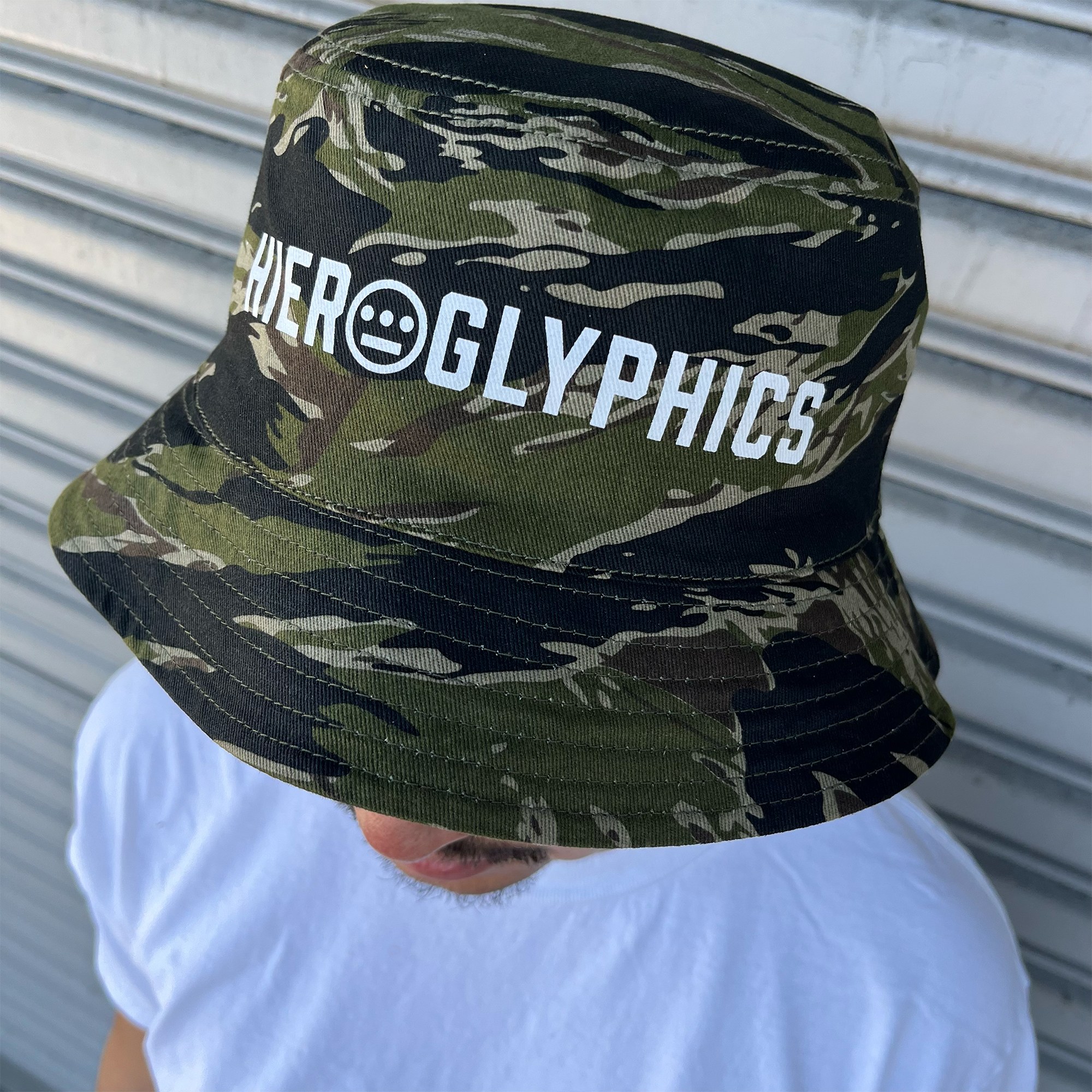 A person wearing the Hiero Reversible Camo Bucket hat by Hieroglyphics, featuring the word "HIEROGLYPHICS" and a smiley face logo on the front, stands in a white t-shirt against a metallic, corrugated surface. The shot is taken from above, with a focus on the tiger camo pattern of the hat.