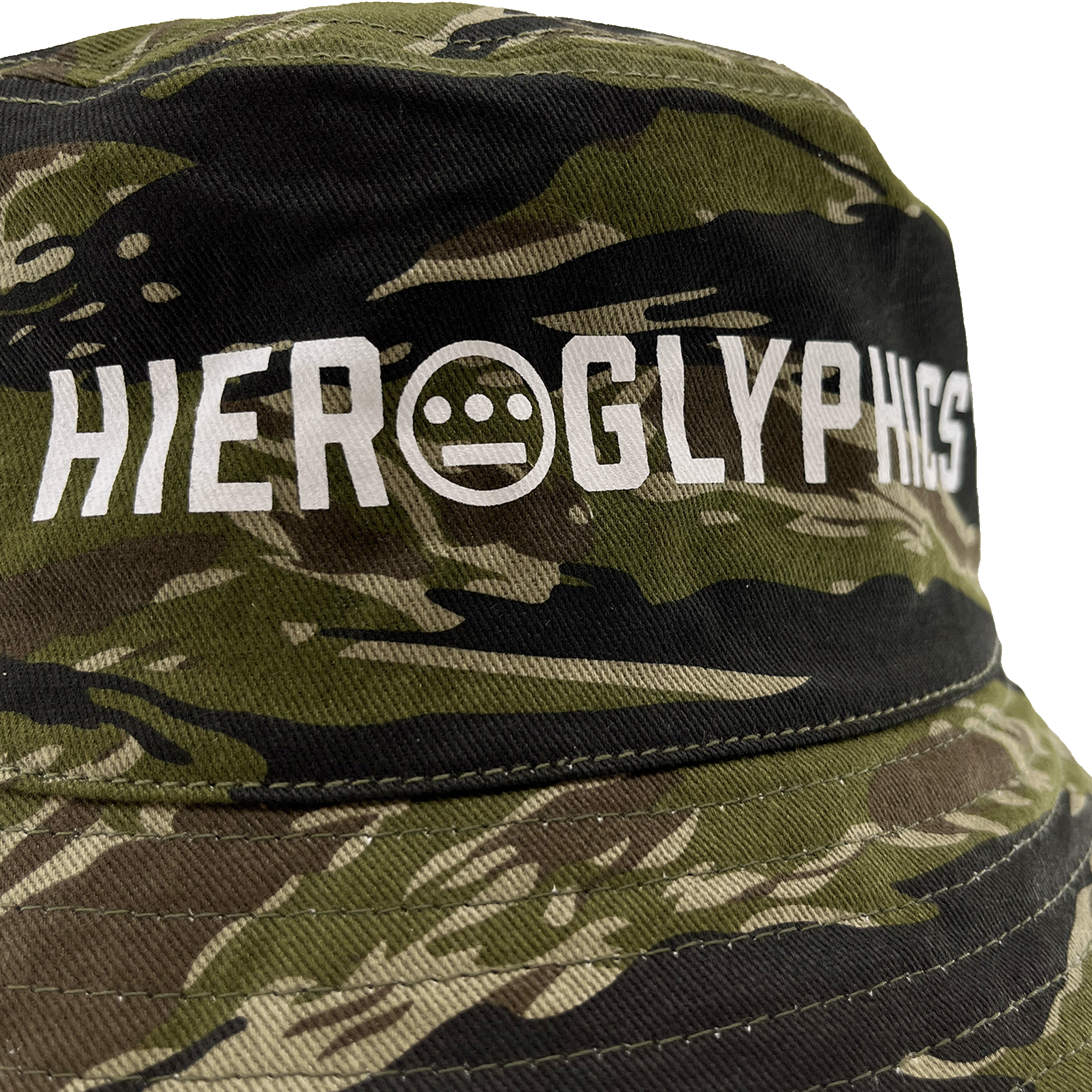 A close-up view of the Hiero Reversible Camo Bucket hat by Hieroglyphics, showcasing the word "Hieroglyphics" in white, uppercase letters. The text features a unique circle design with three horizontal lines and two dots forming a face-like symbol in place of the letter "O.