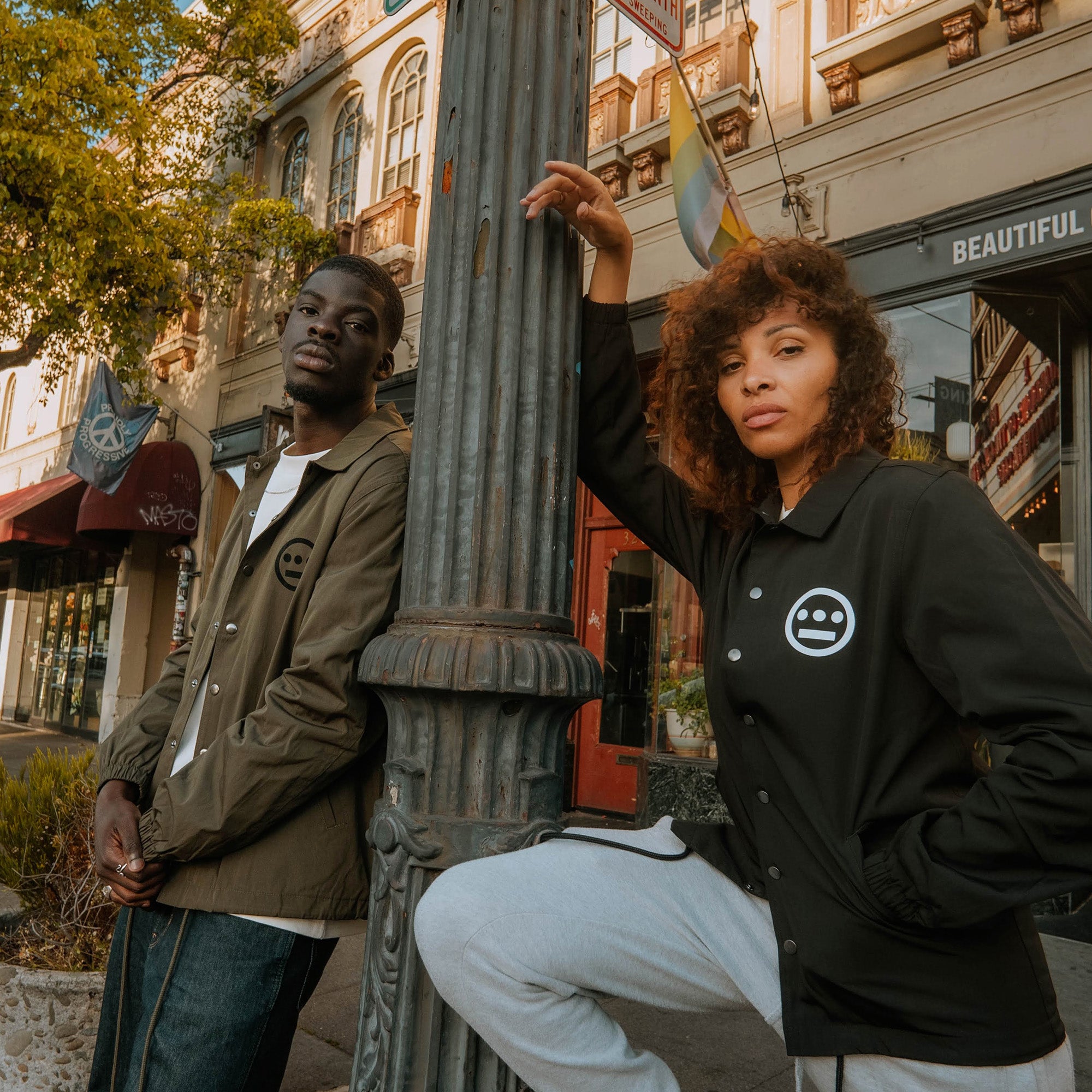 Two people pose confidently on a city sidewalk. The person on the left leans against a lamppost wearing a green, water-resistant Hieroglyphics Premium Coaches Jacket. The person on the right, with curly hair, rests her arm on the lamppost and has a leg raised, wearing a black Hieroglyphics Premium Coaches Jacket featuring a minimalist Hieroglyphics logo.