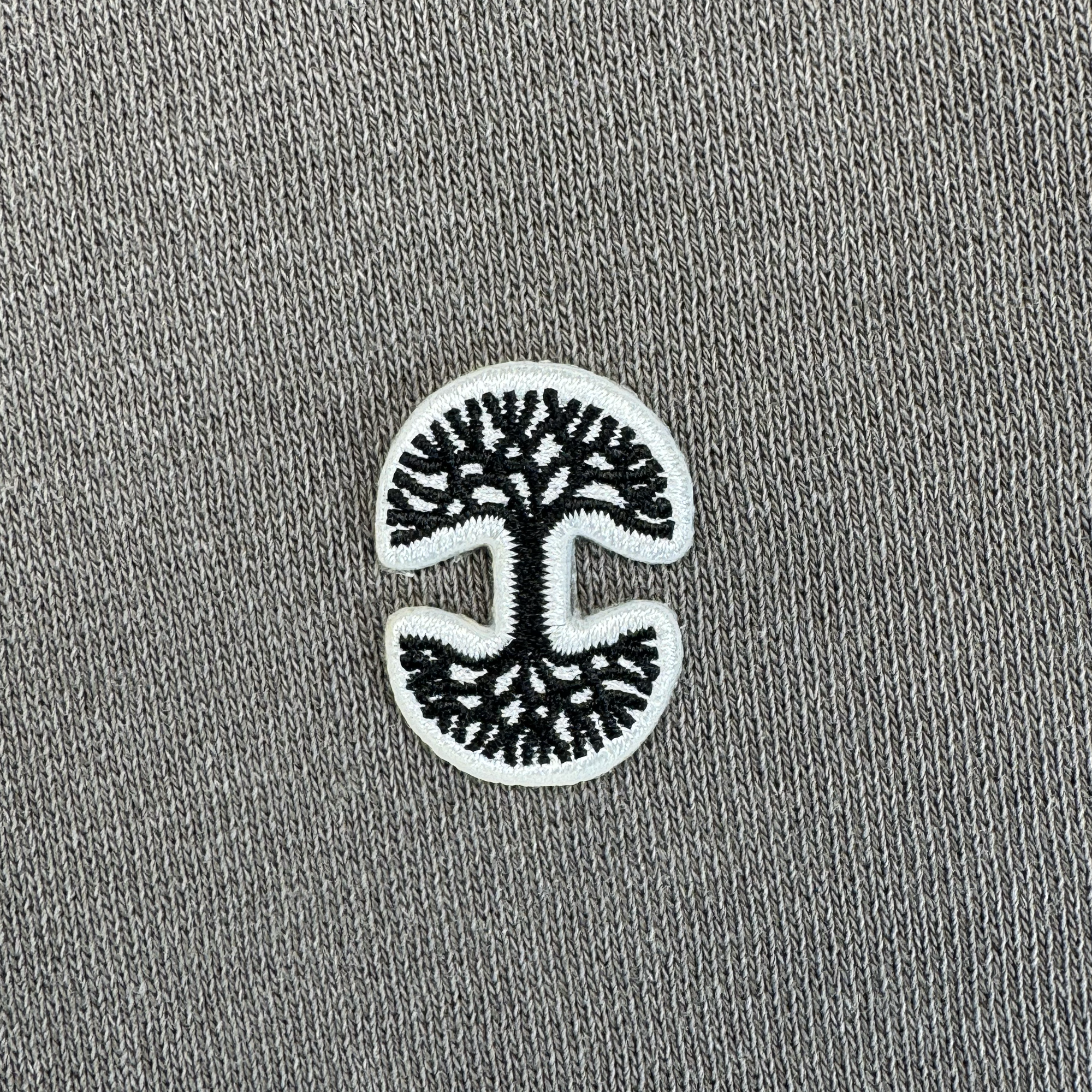 A black embroidered tree with roots and branches is centered in a white oval patch—open on the sides in a “C” shape—on garment-dyed gray Hojicha 1/4 Zip by Oaklandish, perfect for mid-weight outerwear or men’s fleece.