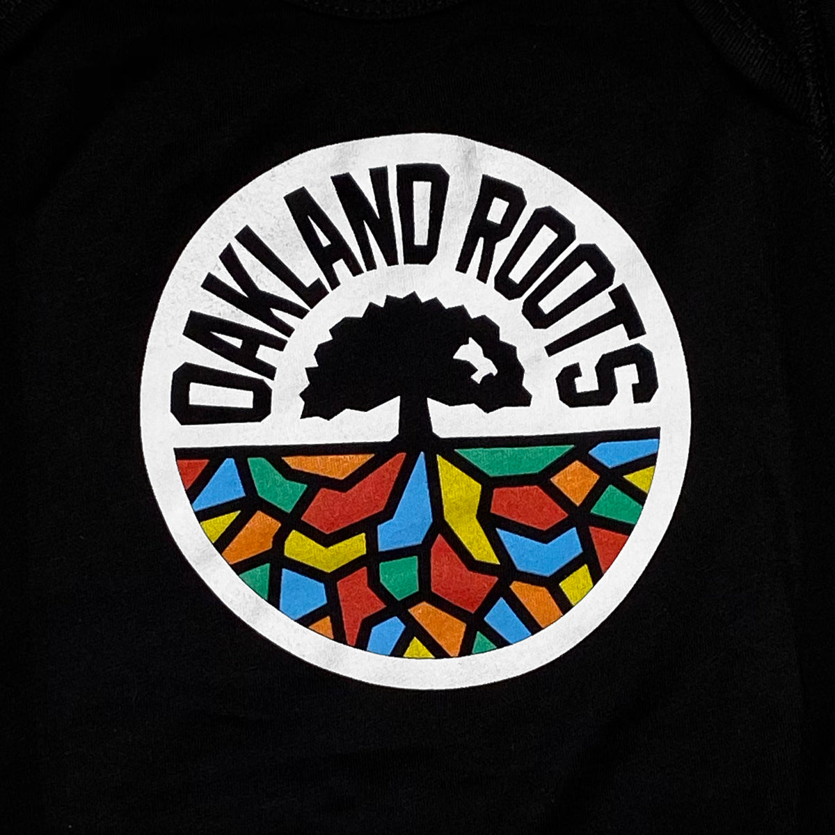 The Infant Oakland Roots SC Logo One-Piece by Oakland Roots SC features a logo design with a white circle, inside of which the words "Oakland Roots" are arched along the top. Below this text is a stylized tree design with black roots extending into a vibrant mosaic of colorful, abstract shapes in red, blue, yellow, green, and orange. Perfect for showing your support for your favorite sports club on an adorable baby romper. The background is black.