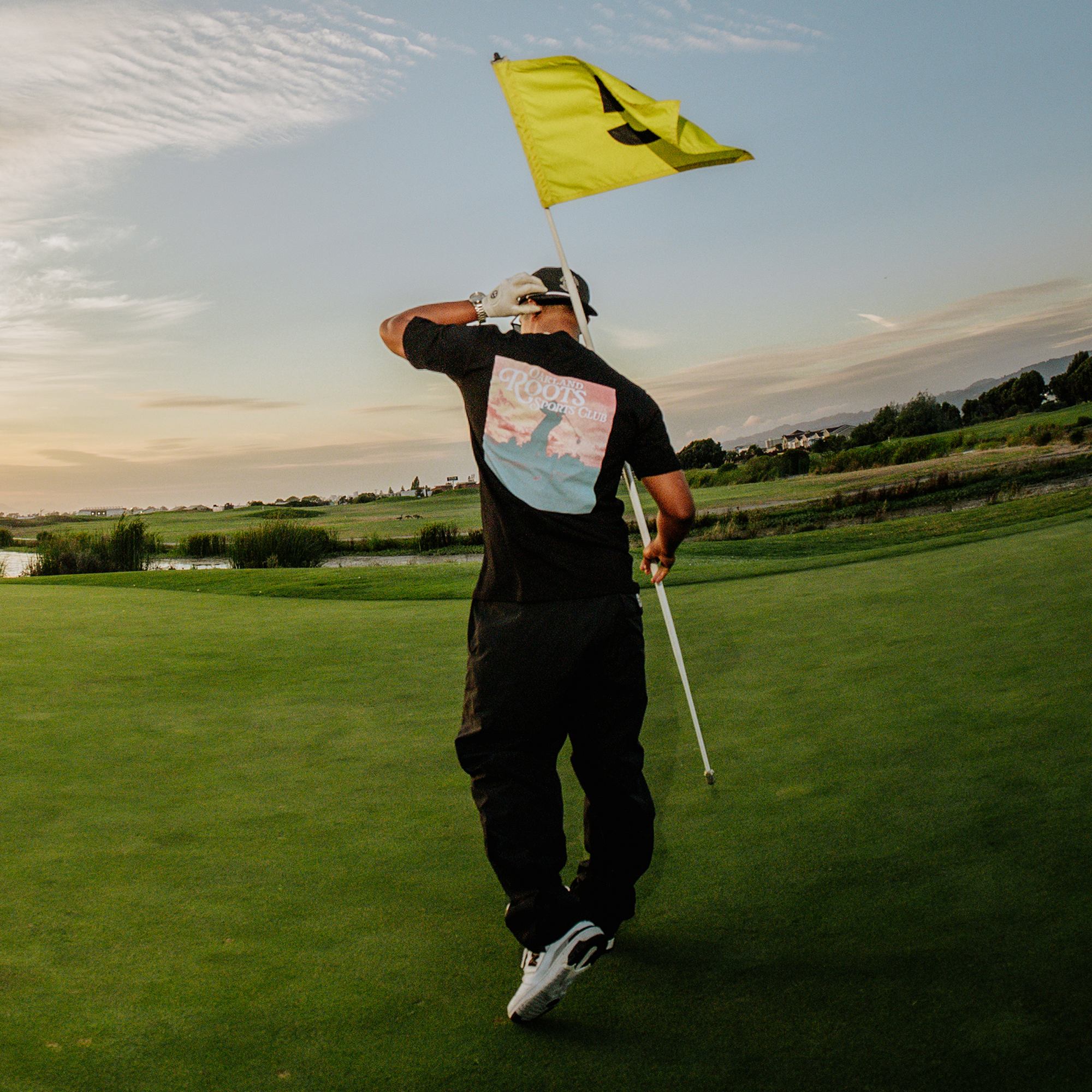 A person in an Oakland Roots SC Legacy Golf Tee, black pants, and white sneakers walks across a green golf course holding a yellow flag with the number "4". The sun is setting, casting a warm glow and creating long shadows. The sky is partly cloudy with hues of orange and pink. Trees and a water body are visible in the background, capturing the serene beauty often appreciated by members of local sports clubs.