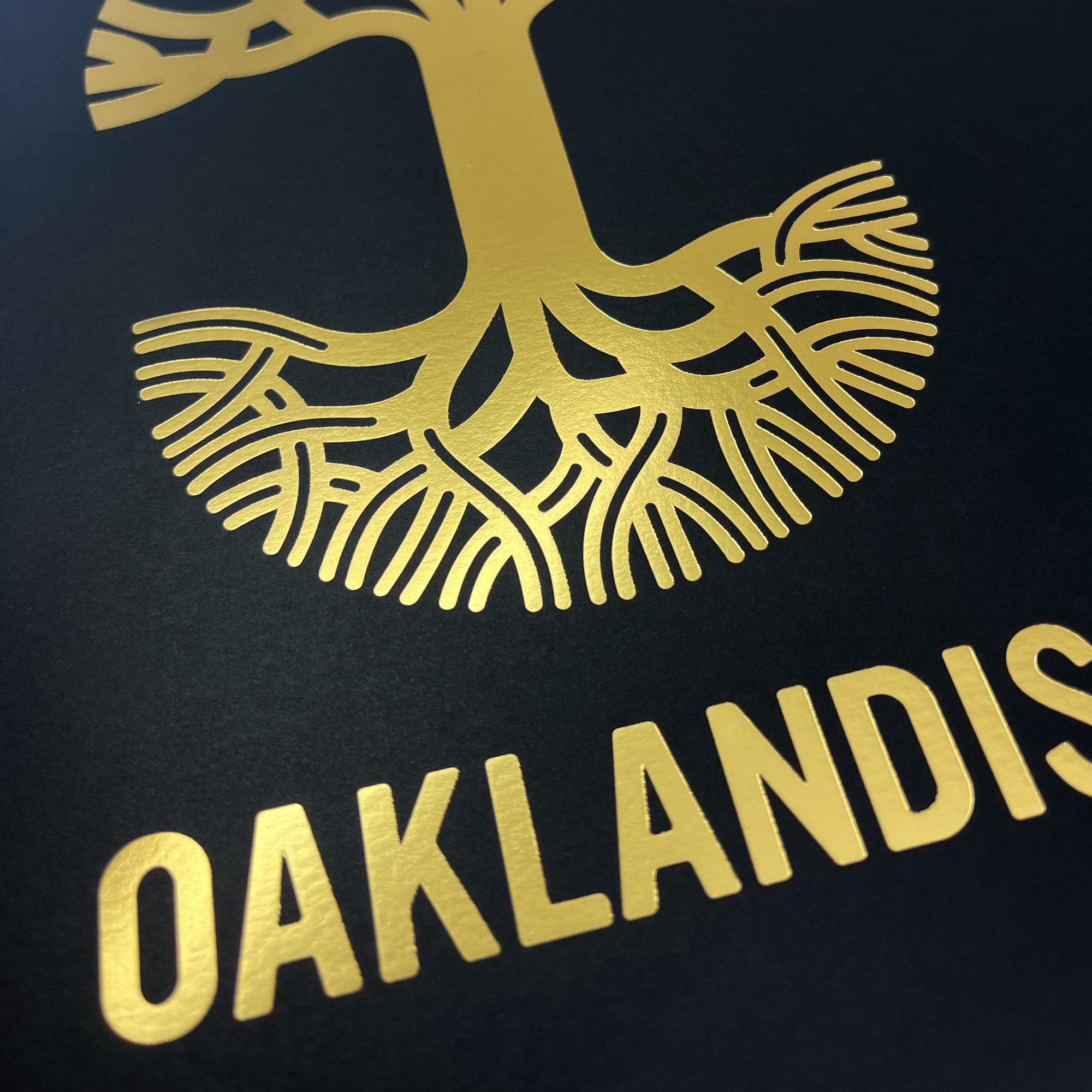 The Oaklandish New Logo Print by Never Elsewhere showcases a striking gold tree with intricate roots set against a dark background. Beneath the tree, "OAKLANDISH" is emblazoned in bold, copper foil letters. The design creates a stunning contrast between the shimmering gold and the dark backdrop, evoking the sophistication of an elegant letterpress on fine cardstock.