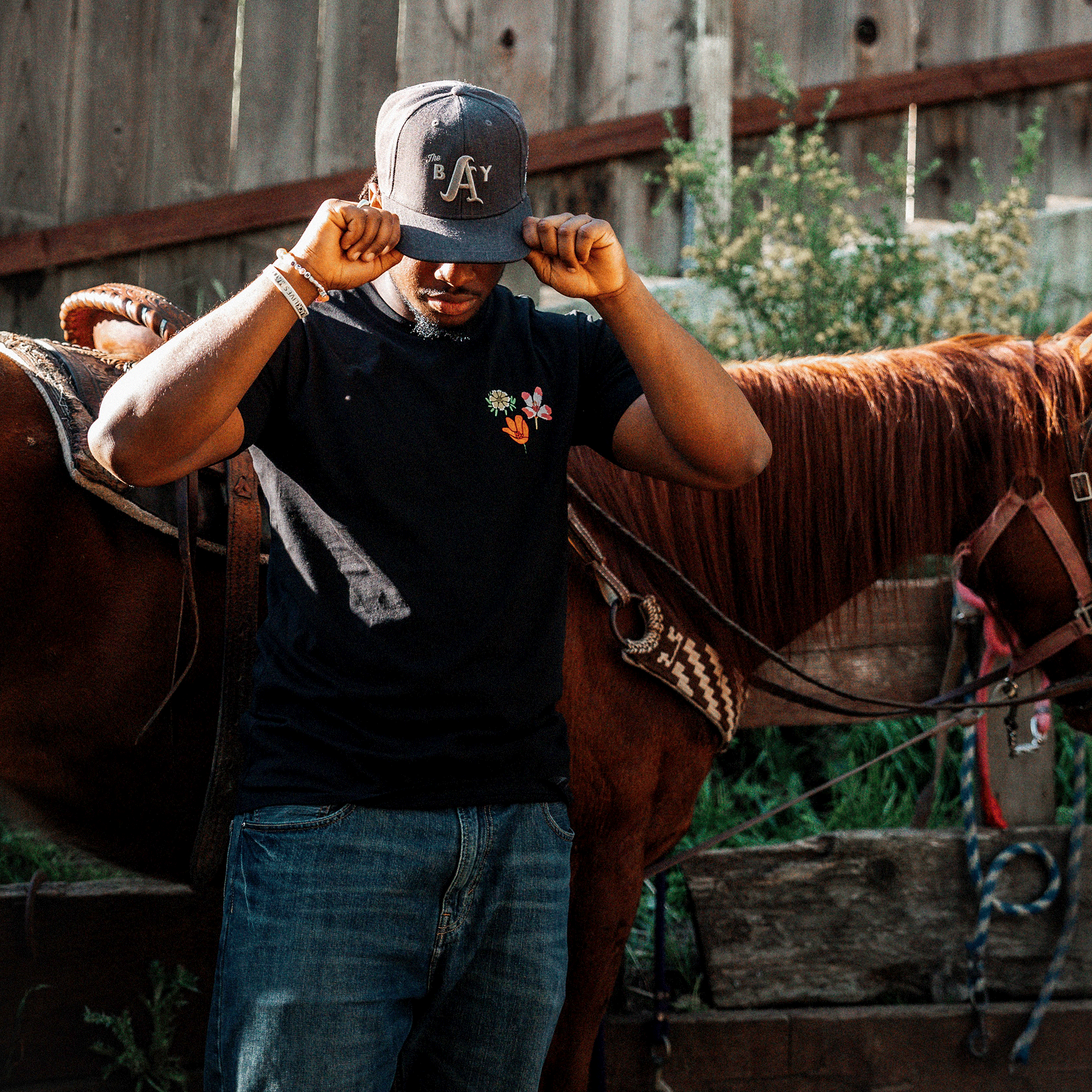 A person wearing a dark cap, an Oakland Wildflowers classic fit black t-shirt from the Oaklandish men's collection, and blue jeans stands in front of a brown horse saddled with traditional gear. The person is adjusting their cap with both hands. The background features a wooden fence and foliage, providing a rustic outdoor setting.