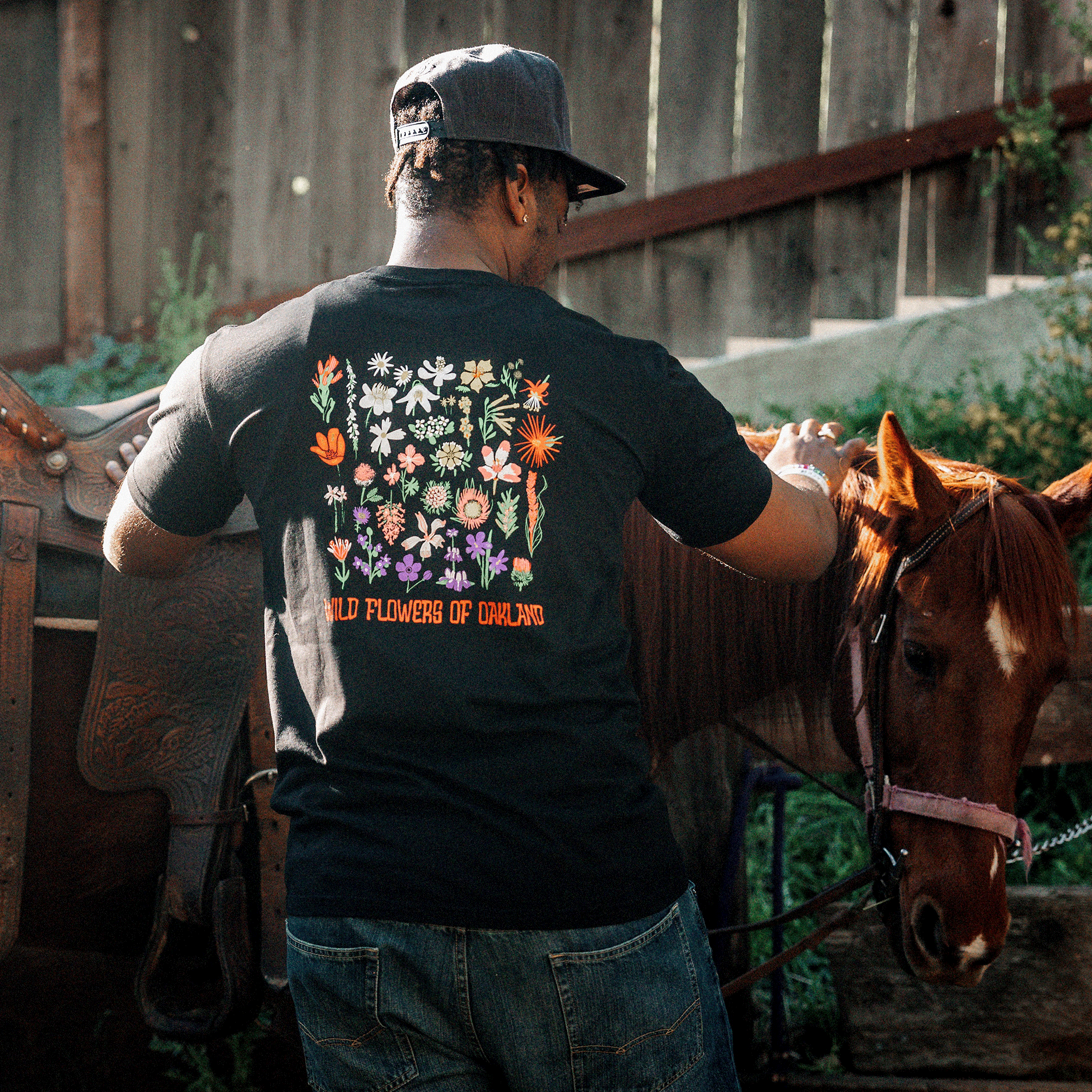 A man wearing a black cap and an Oaklandish "Oakland Wildflowers" classic fit T-shirt with colorful wildflowers and the text "Wild Flowers of Oakland" printed on the back leans over a brown horse, gently holding its reins. The horse is equipped with a saddle. The background includes a wooden fence and green foliage.