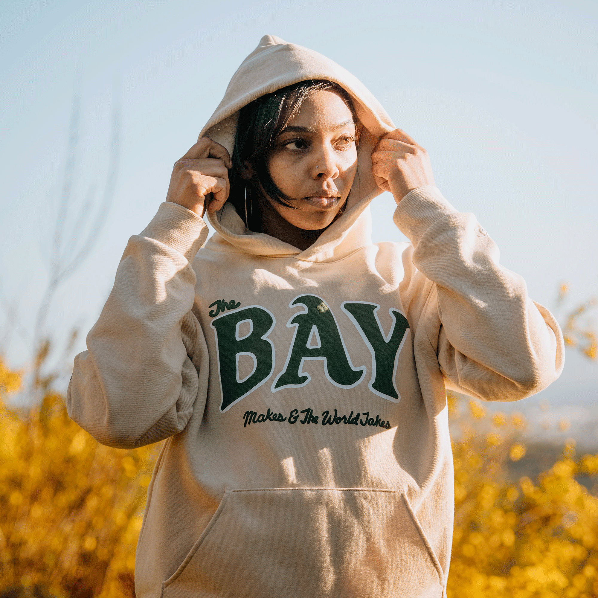 A person stands outside in golden sunlight, adjusting their beige "The Bay Hoodie by DOC" from Oaklandish, crafted from premium heavyweight fleece. This hoodie is part of a capsule collection celebrating Bay Area culture and showcases "The Bay" in large green letters with "Make & The World Takes" underneath. They are surrounded by yellow foliage and a clear blue sky.