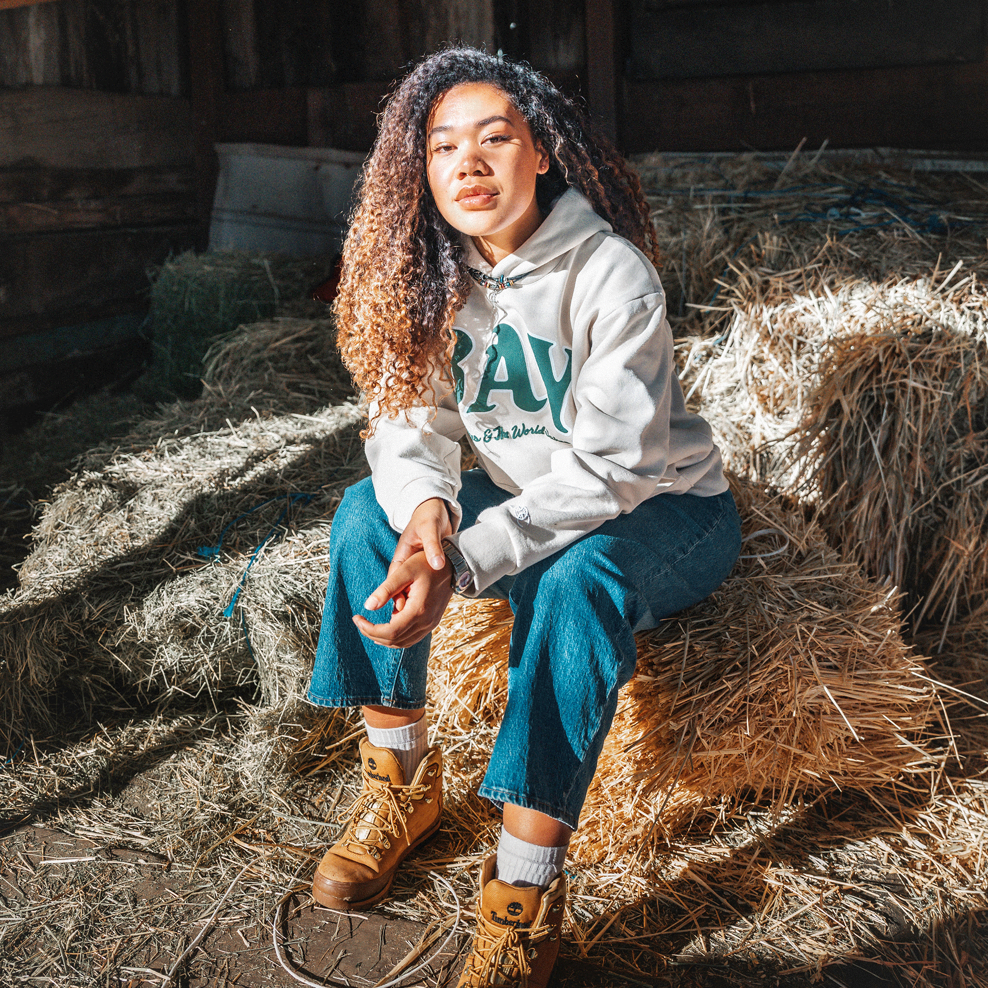 A woman with curly hair, wearing The Bay Hoodie by DOC from Oaklandish's capsule collection—a light-colored hoodie with green lettering—paired with blue jeans and tan boots, sits on a pile of hay in a rustic wooden barn. Sunlight streams in, casting shadows on the floor and around her. She looks directly at the camera with a neutral expression.