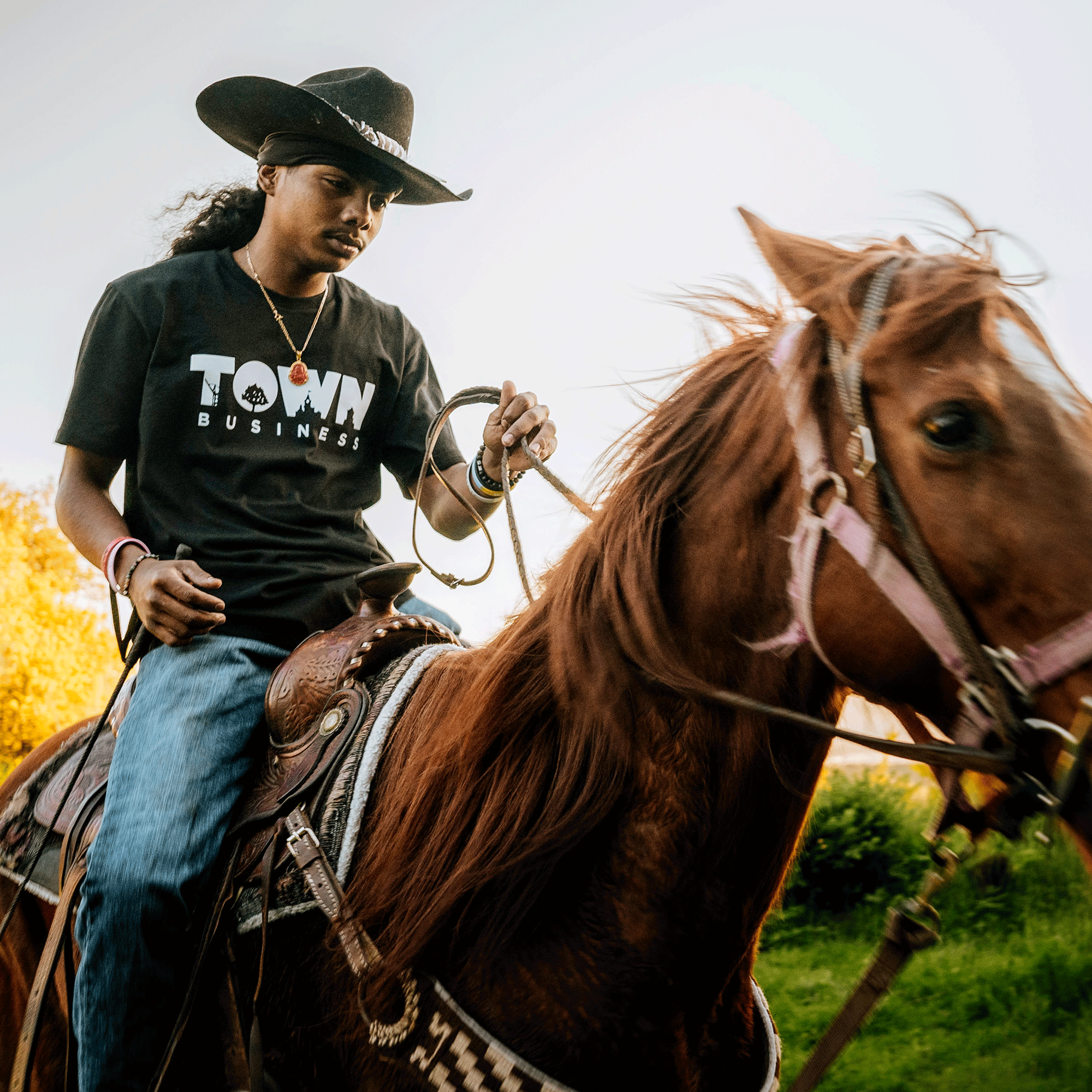 A person with long hair, wearing a black cowboy hat and an Oaklandish "Town Business 2024" Tee, is riding a brown horse. The rider holds the horse's reins while looking ahead. The background features East Bay greenery and sunlight filtering through the trees.