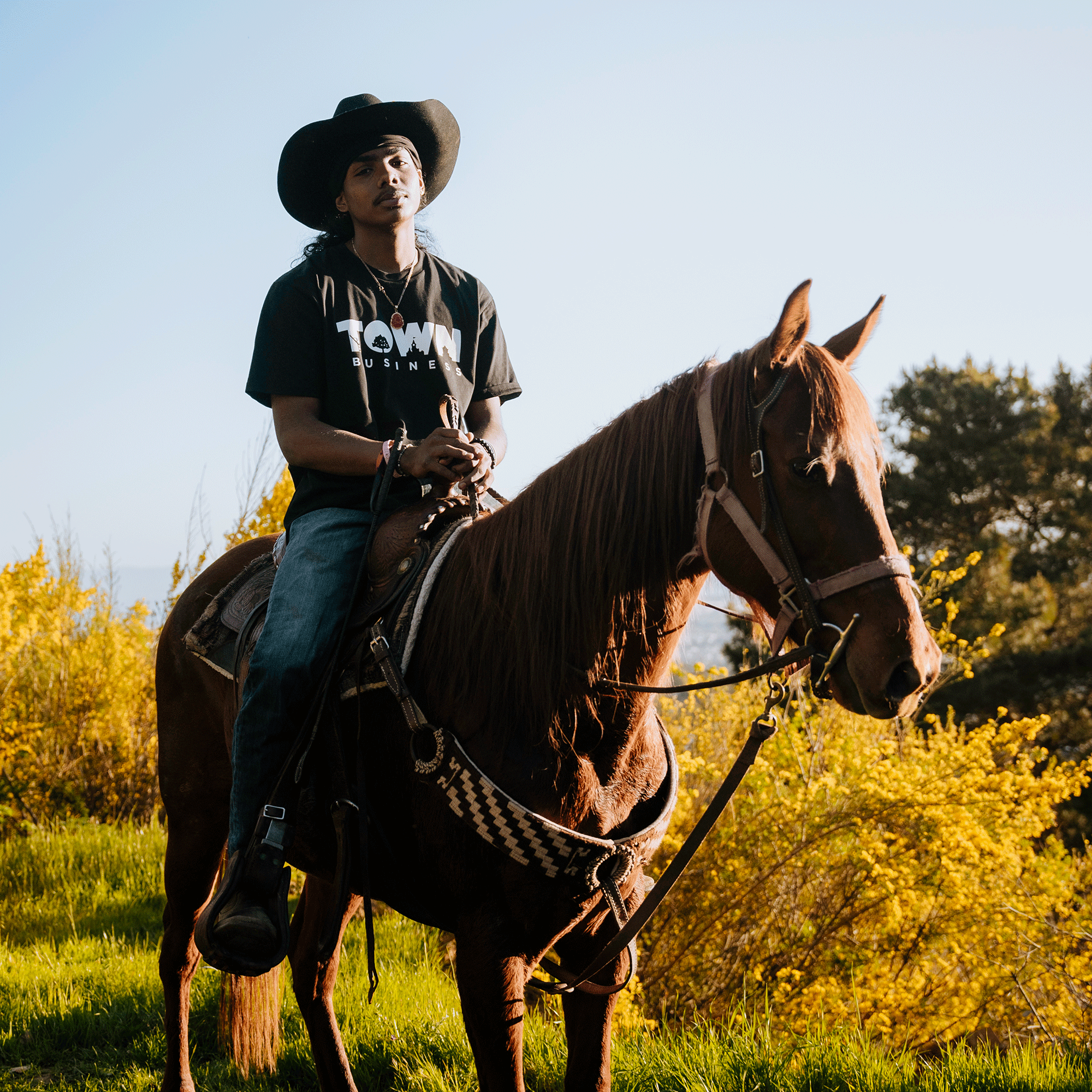 A person wearing a black cowboy hat, an Oaklandish Town Business 2024 Tee in black, and blue jeans is seated on a brown horse in the East Bay. The horse has a bridle and saddle, and they are outdoors in a grassy area with yellow flowers and trees in the background, under clear blue skies.