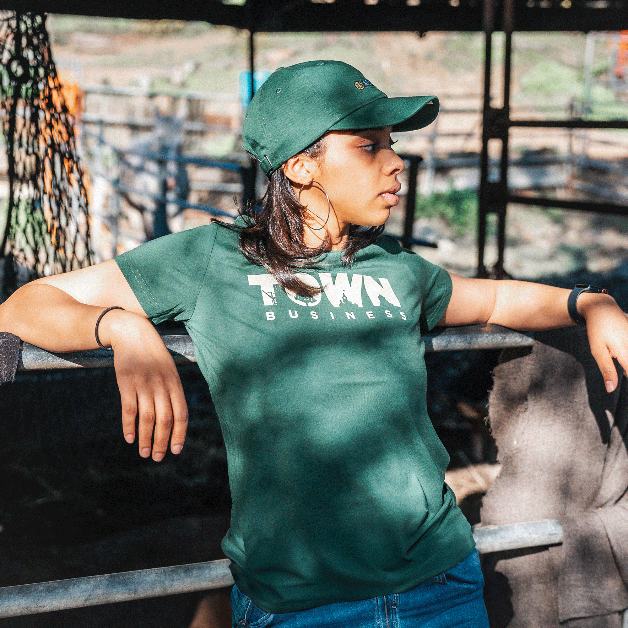 A woman in a green "Women's Town Business 2024 Tee" by Oaklandish and a cap leans against a metal railing outdoors. Her right arm rests on the railing, while her left hand touches it lightly. The background shows a blurry outdoor scene with what appears to be a farm structure and some greenery.