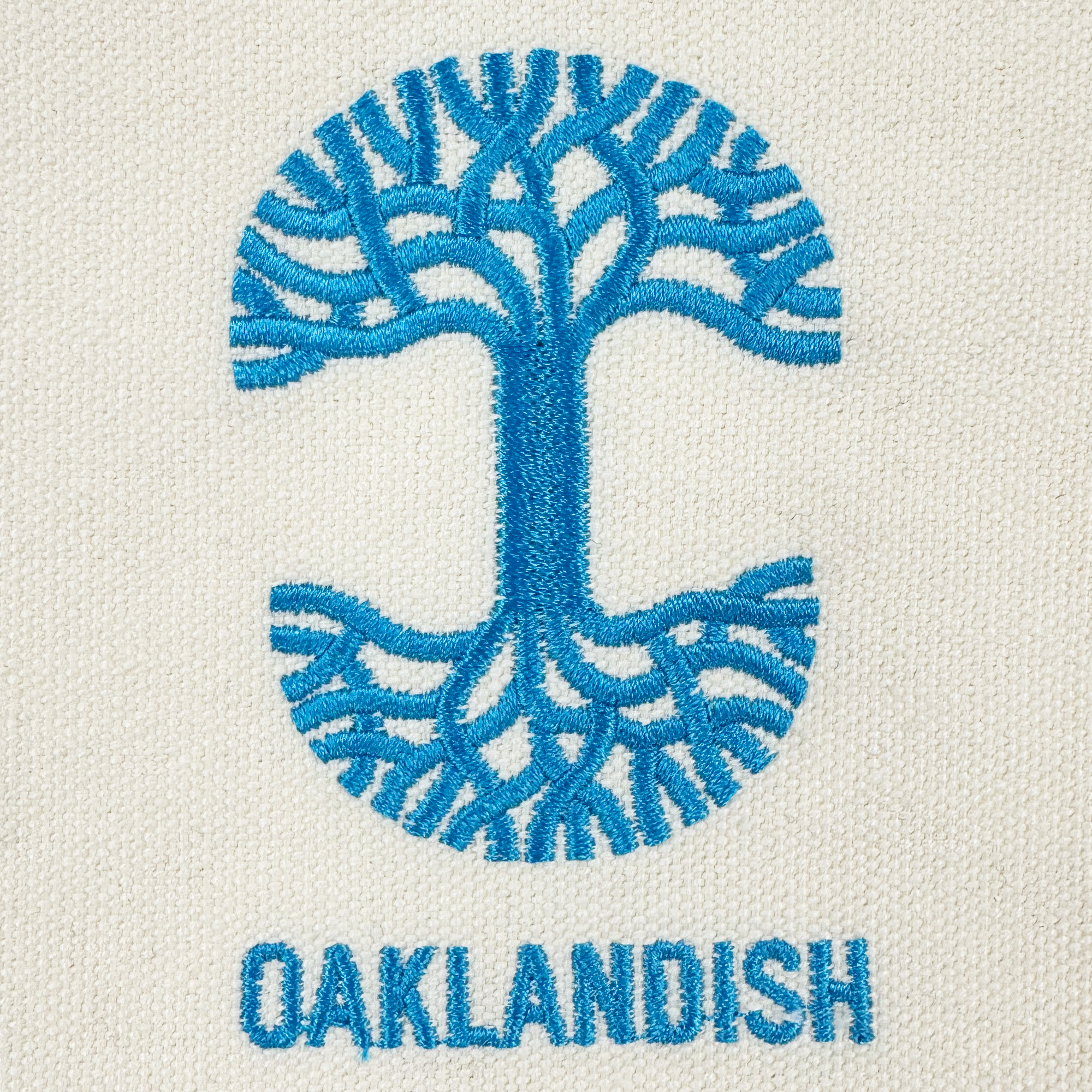 The Oaklandish Mini Grocery Tote features a blue embroidered tree logo with branching roots and limbs in a circle above "OAKLANDISH" on cream-colored fabric—perfect as an accessory or statement piece from the Oaklandish brand.