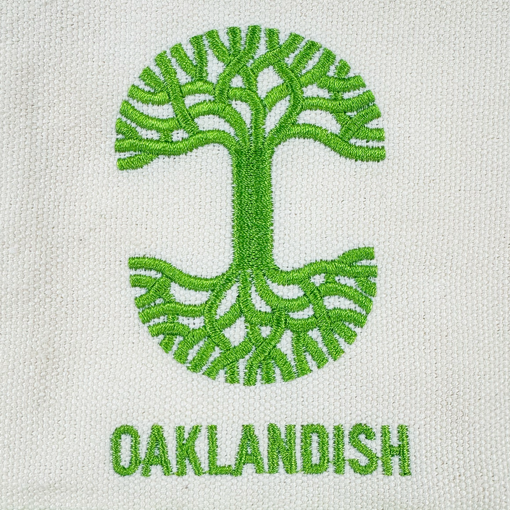 A green oak tree with spreading branches and roots forms an oval above the lime green "OAKLANDISH" logo, all on white textured fabric—featured on the Oaklandish Mini Grocery Tote by Oaklandish, ideal for stylish everyday use.