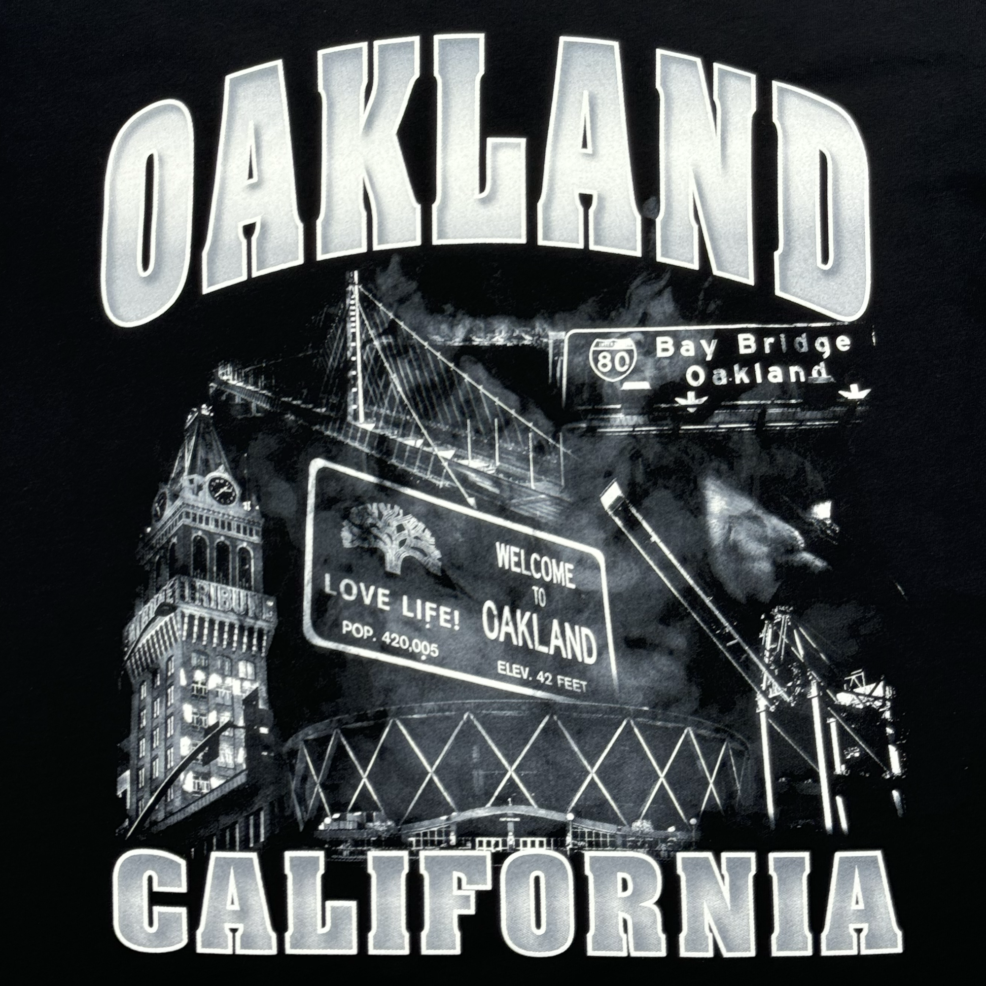 A montage design presents "OAKLAND CALIFORNIA" in bold silver letters, featuring the Bay Bridge, a "Welcome to Oakland" sign complete with population and elevation details, as well as local landmarks. It's all styled on a classic fit t-shirt from Oaklandish's Montage Tee line against a dark background.