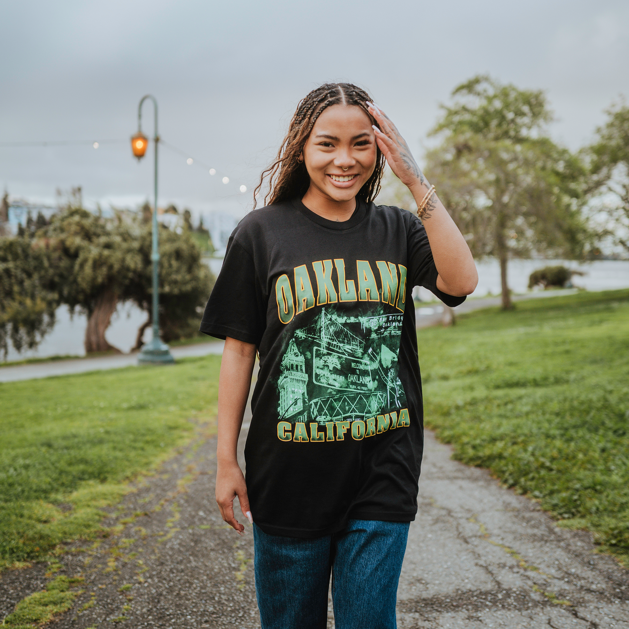 A smiling woman with long braided hair, wearing an Oaklandish Montage Tee and jeans, walks through a park. As she touches her hair, the grassy area and large tree evoke Emerald City vibes under a clouded sky with lights strung from a lamppost.
